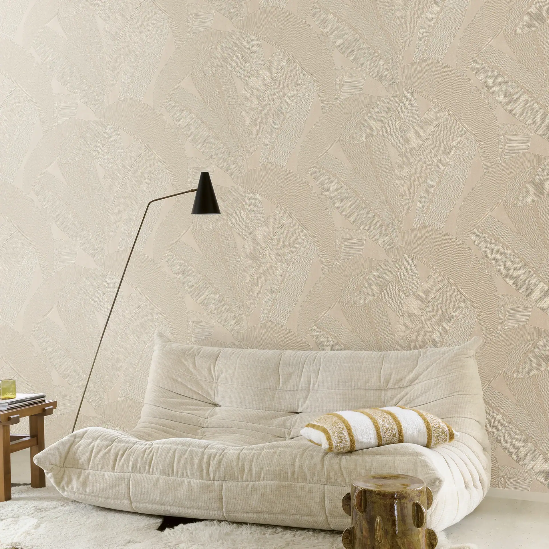 A neutral-toned living room with a beige modular sofa, a black floor lamp, and light beige wallpaper patterned with large, stylized banana leaves.