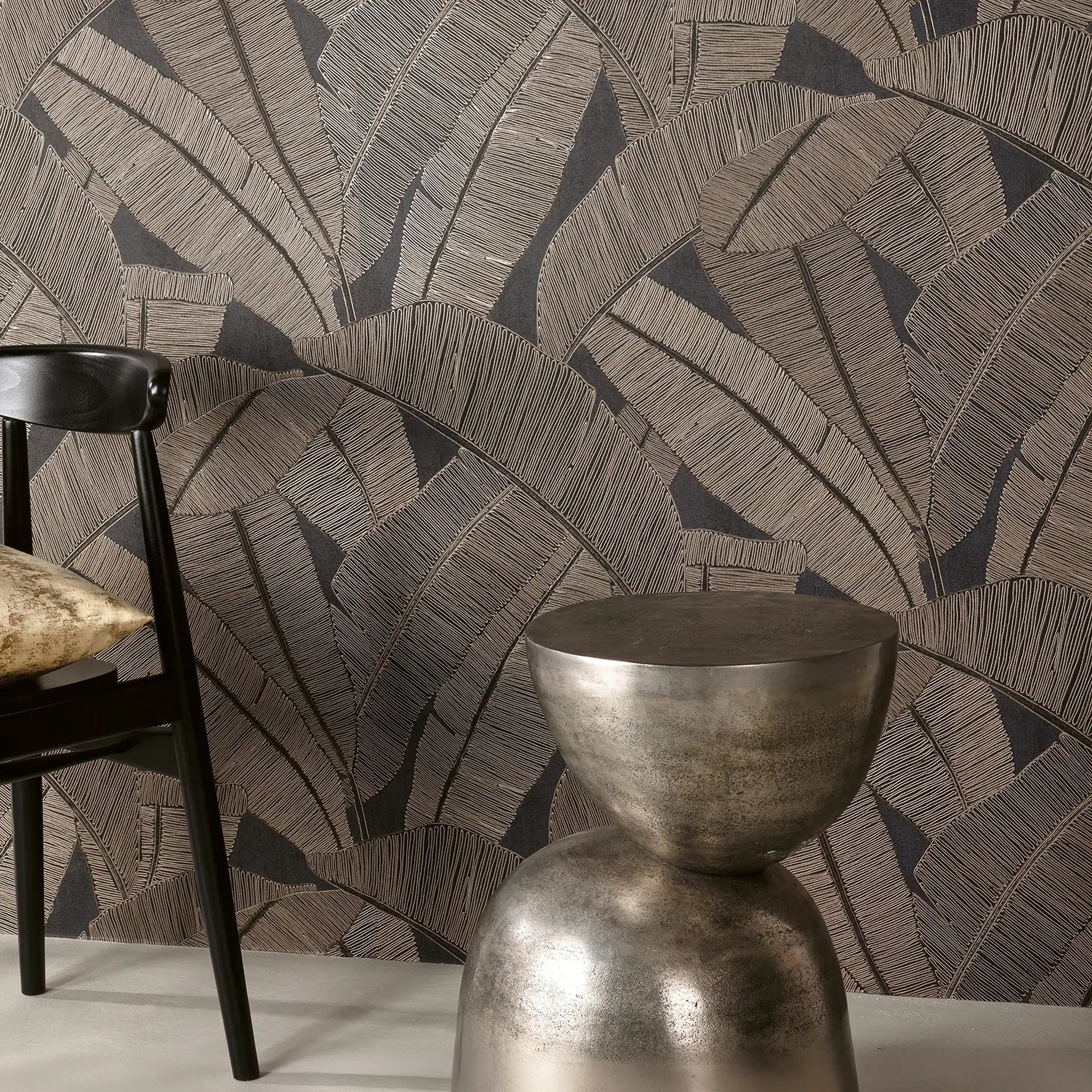 A dark wallpaper with a textured, light bronze leaf pattern. A black chair with a gold cushion and a silver metallic side table are in the foreground.