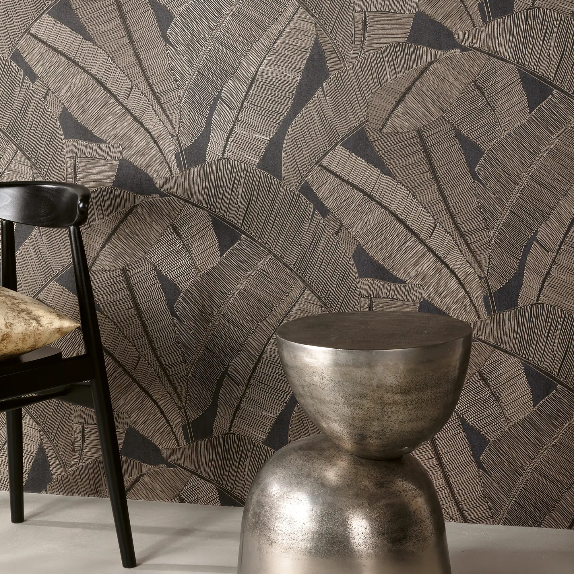A dark wallpaper with a textured, light bronze leaf pattern. A black chair with a gold cushion and a silver metallic side table are in the foreground.