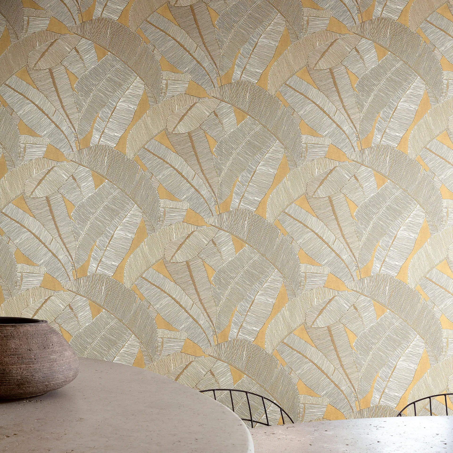 A wall covered in light beige and golden-yellow banana leaf patterned wallpaper, with a light-colored round table, a rustic brown pot, and part of a black metal chair in the foreground.