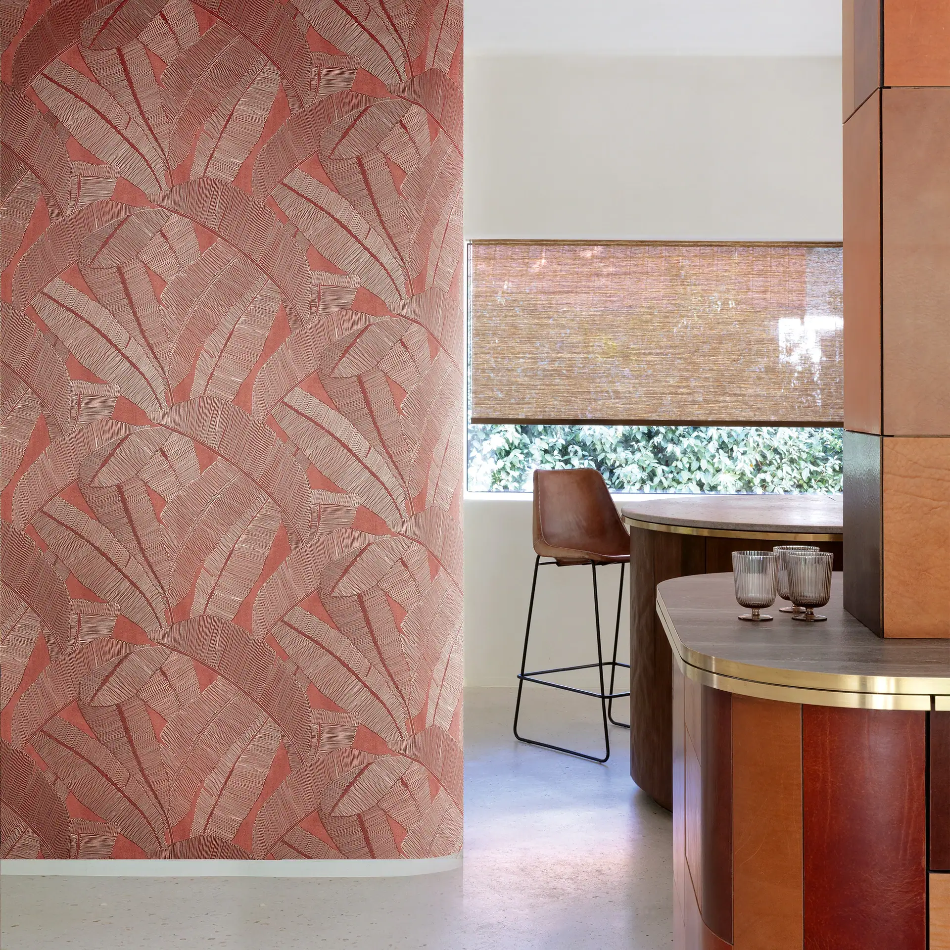 A modern room with red leaf-patterned wallpaper, a curved wooden bar with brass trim, a brown leather stool, and a window with a woven blind.