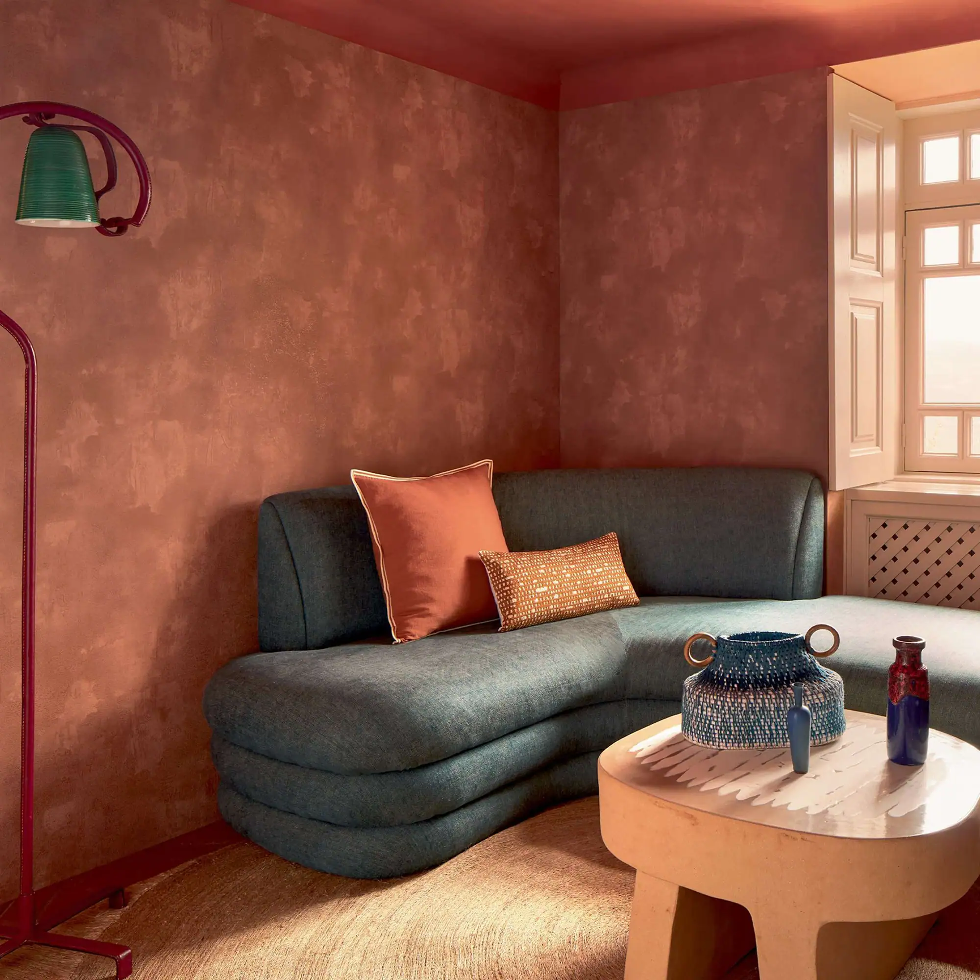 A living room corner with terracotta walls, a dark teal L-shaped sofa, a beige coffee table with vases, and a red floor lamp with a green shade. A white window is on the right.