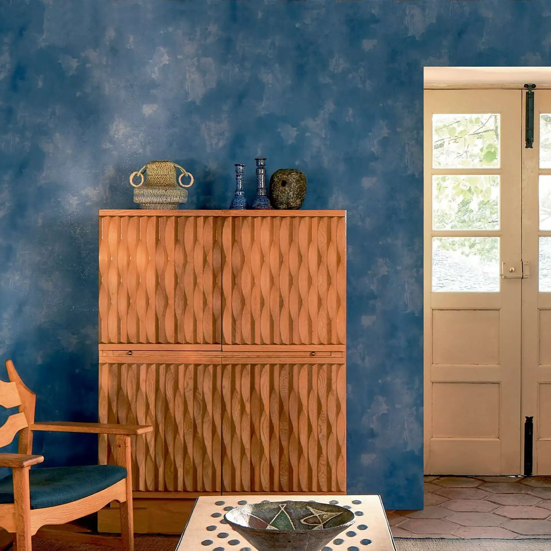 A room with a textured blue wall, a carved wooden cabinet, an armchair, and a doorway with glass panes leading to an outdoor view.