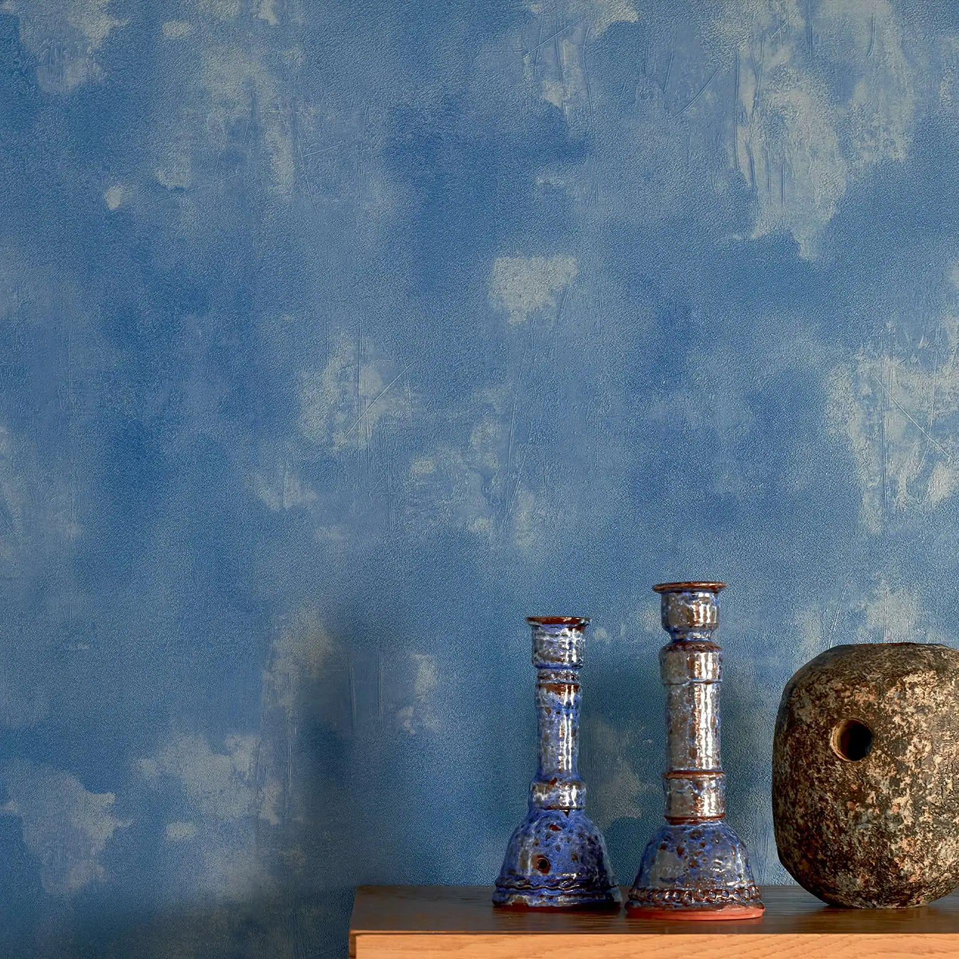 Two blue and brown ceramic candle holders and a dark, speckled stone-like object with a hole, sitting on a wooden surface against a textured blue wall.