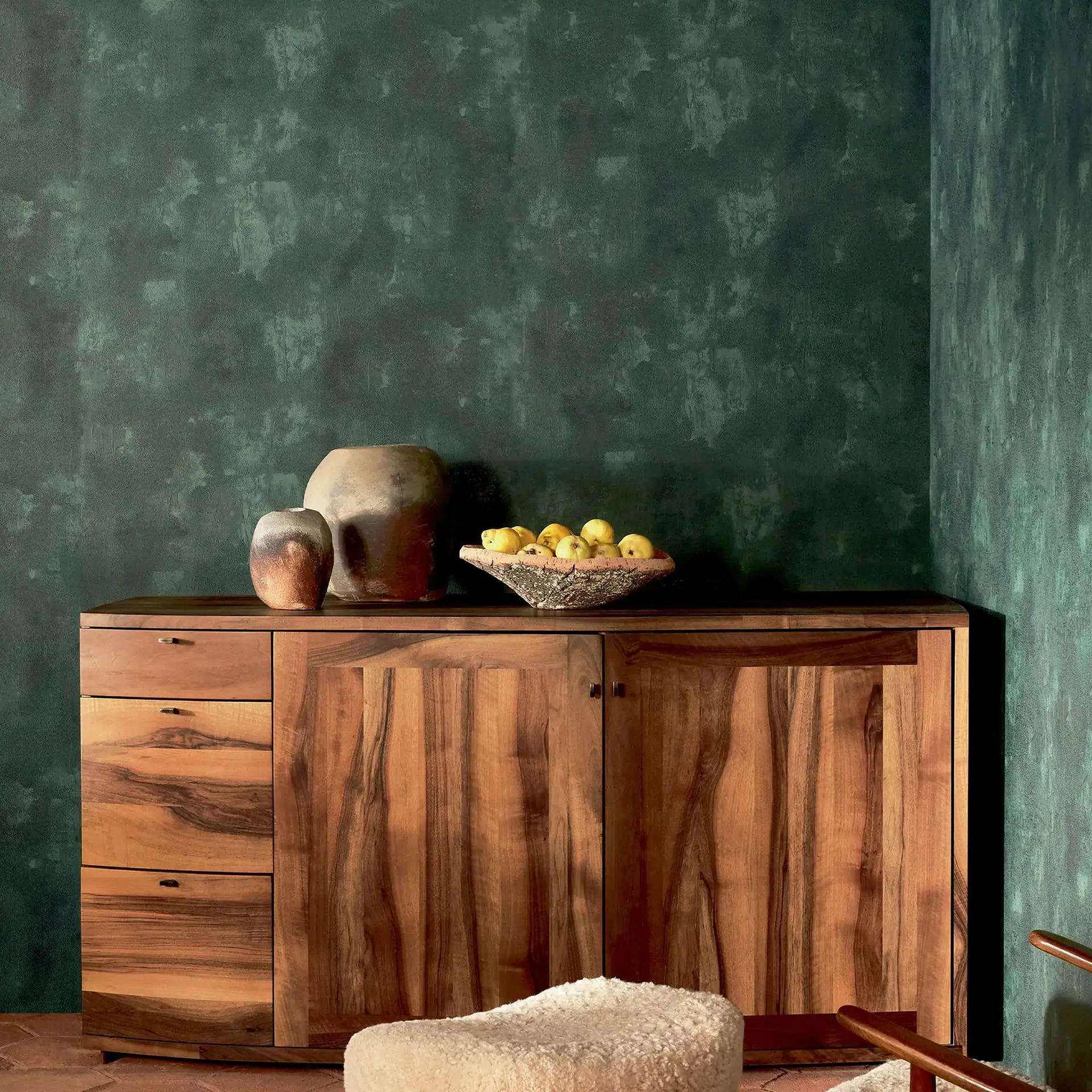 A dark wood sideboard with ceramic vases and a bowl of yellow fruit sits against a textured, mottled dark green wall. A fluffy cream ottoman is in the foreground.