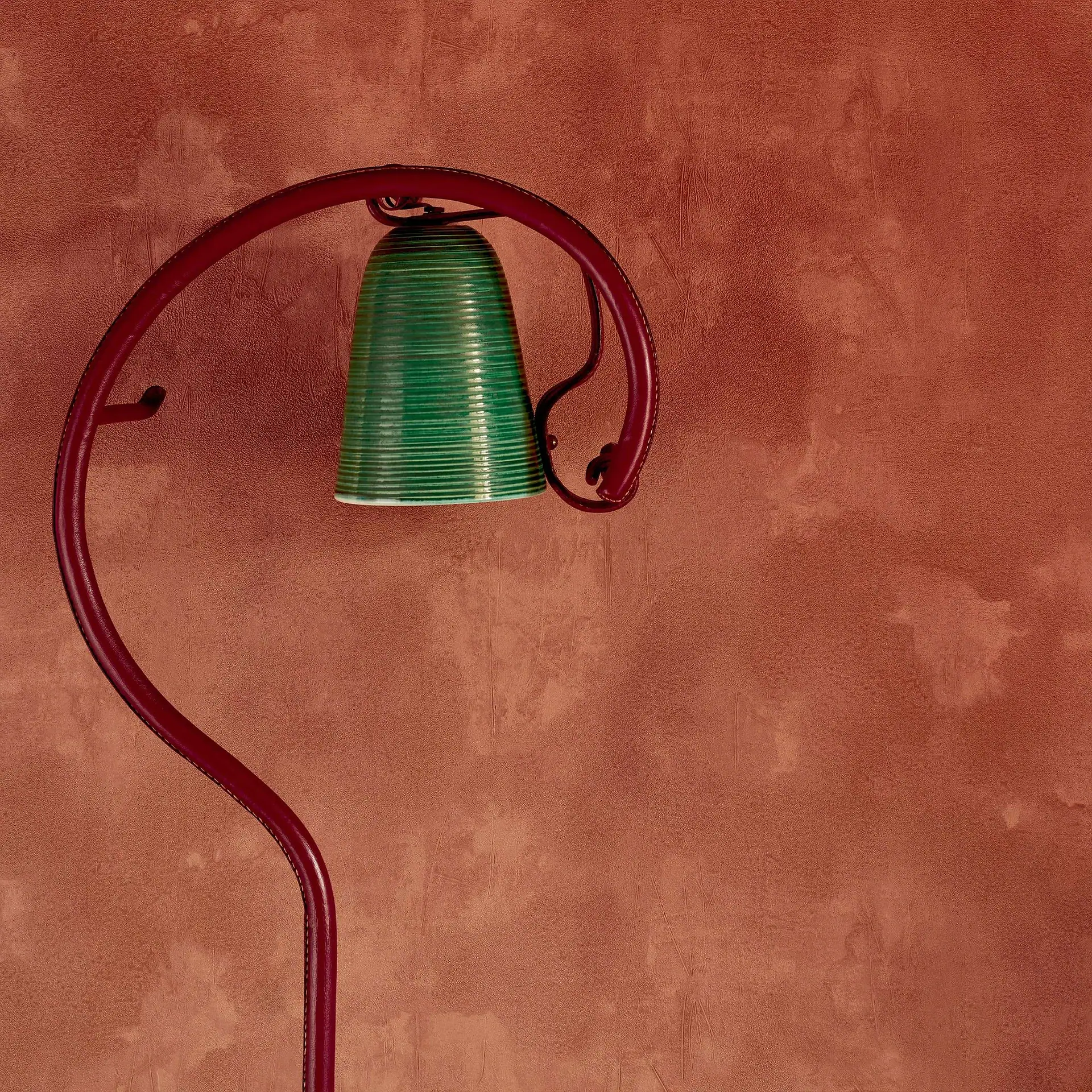 A green ribbed bell hangs from a curved red stand against a textured reddish-brown wall.