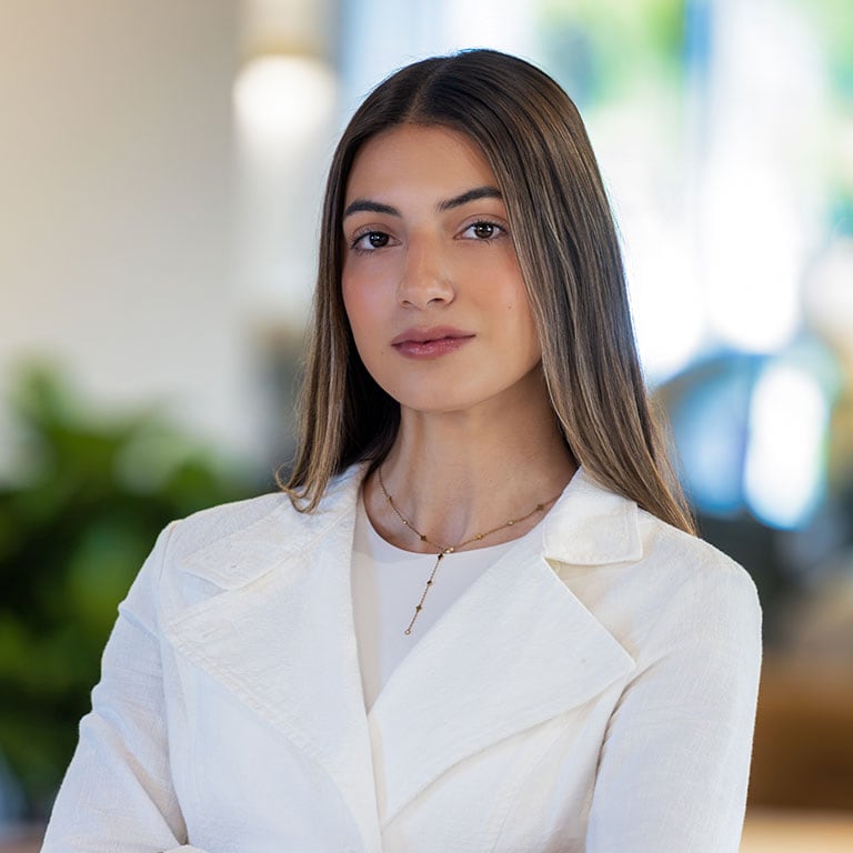 A young woman with long brown hair, wearing a white blazer and a delicate gold necklace, looks directly at the camera.