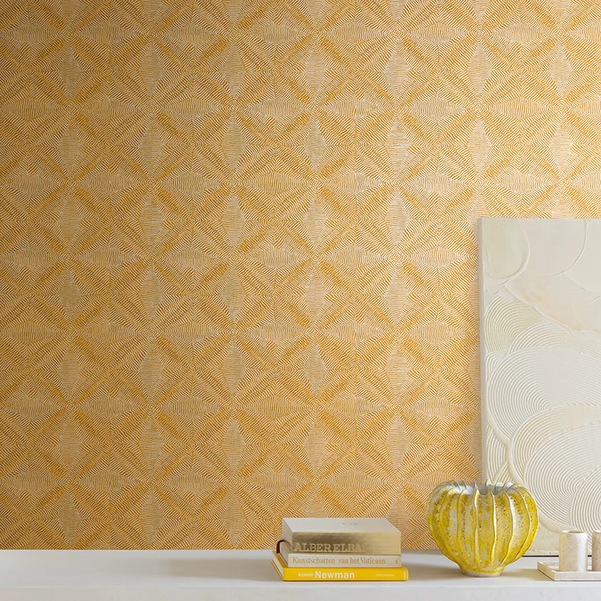 A wall covered in golden yellow geometric textured wallpaper, with a white shelf holding a stack of books, a yellow ribbed vase, and a white textured art piece.