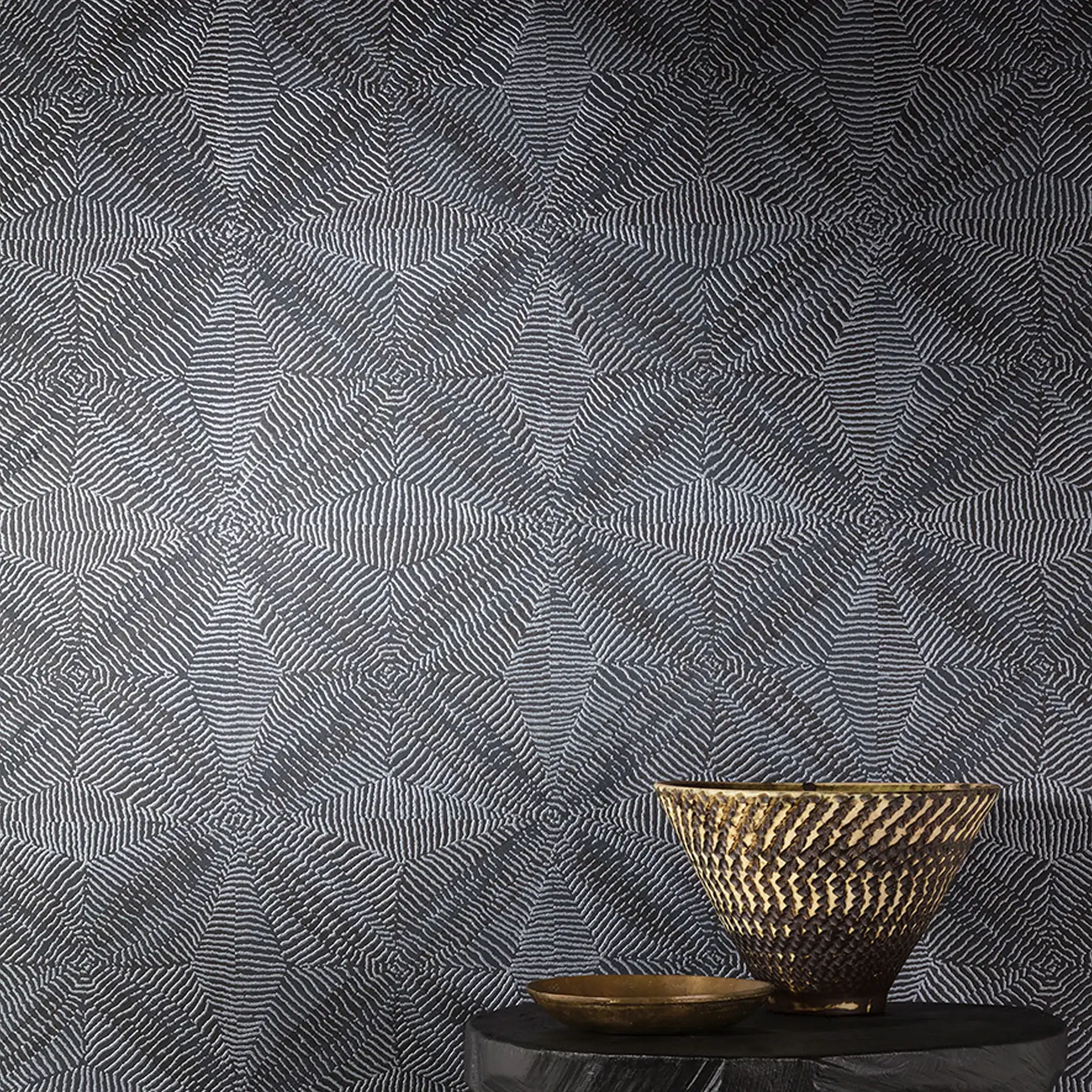 A dark gray wallpaper with a repeating pattern of light gray wavy lines forming concentric squares and diamonds. On a dark table in the foreground, a large textured bronze bowl and a smaller smooth bronze dish are displayed.