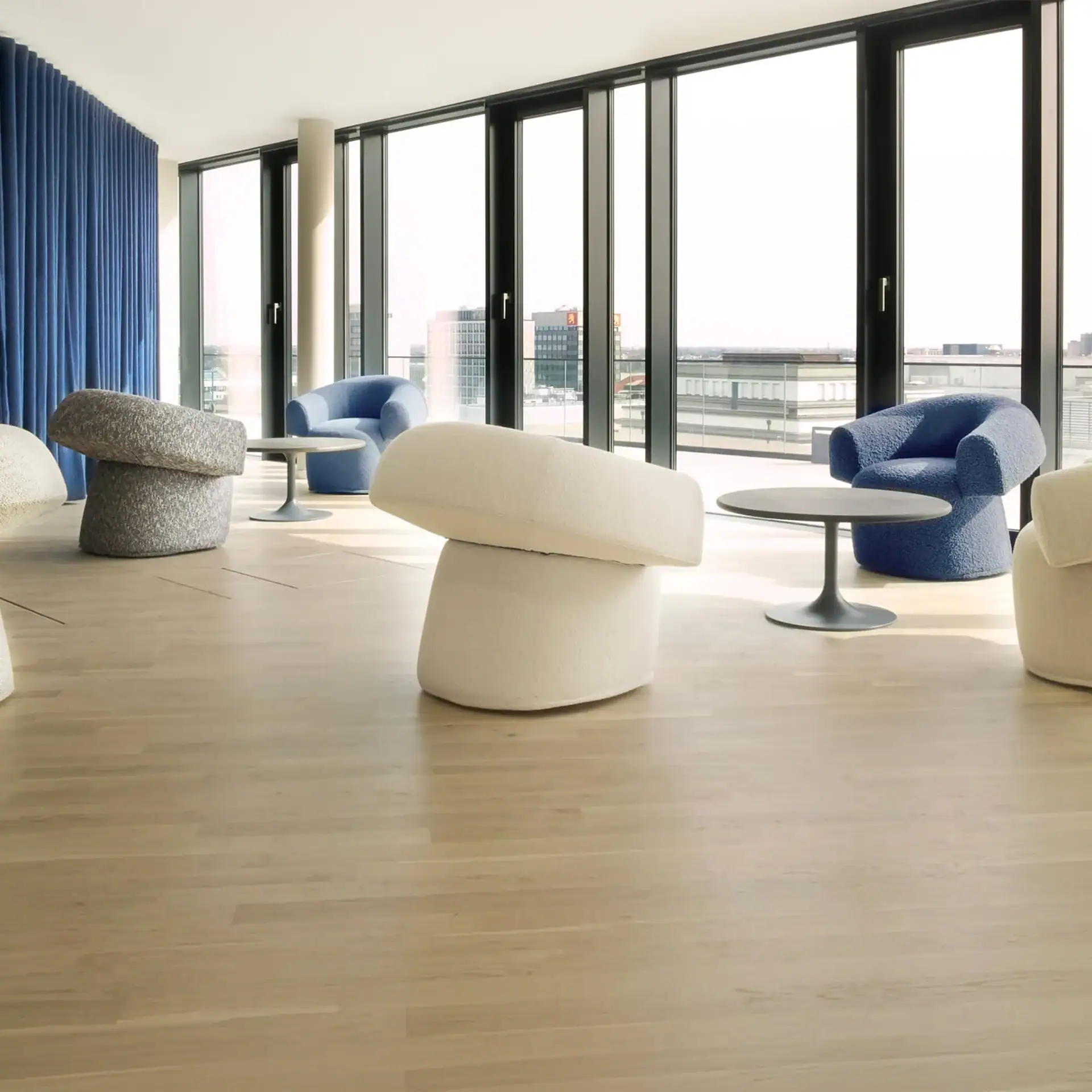 A modern lounge with large windows overlooking a city, furnished with blue and off-white bouclé chairs, stacked cushion ottomans, and round tables.