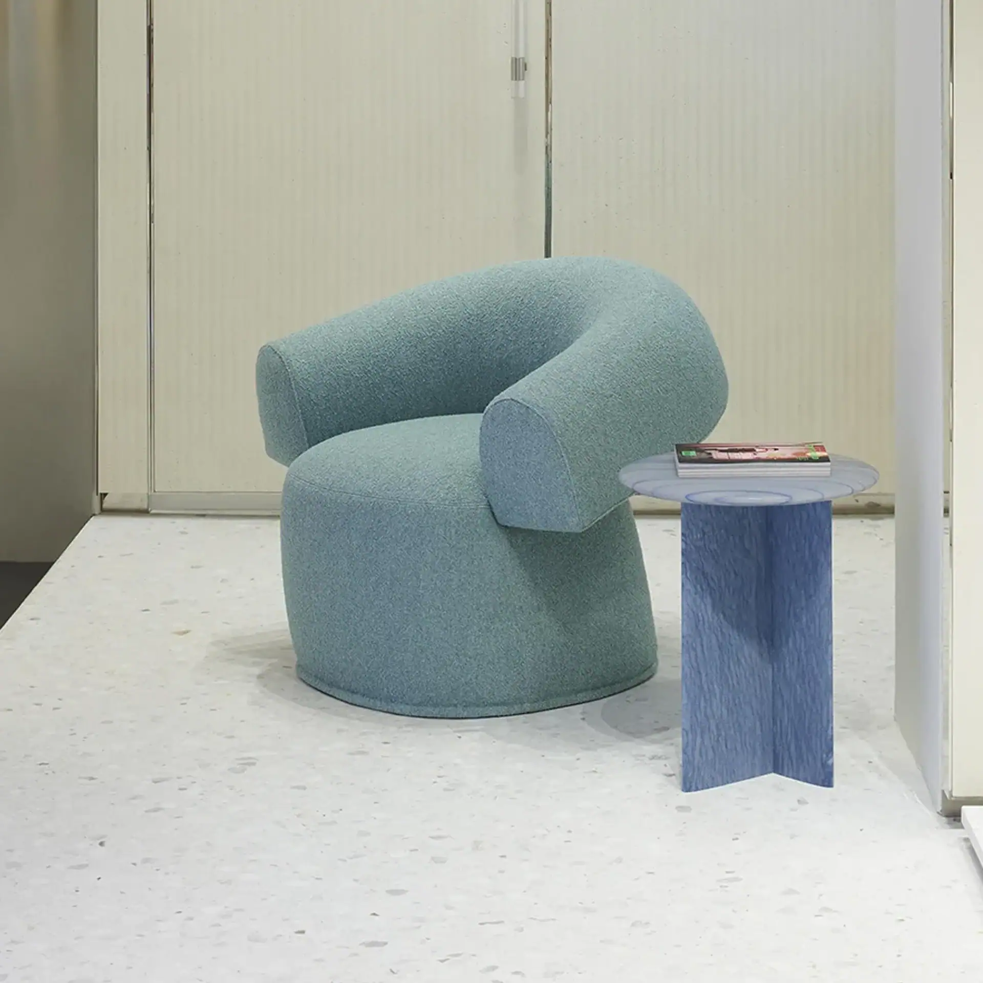 A light blue, rounded armchair next to a blue side table with magazines on a terrazzo floor in a modern room.