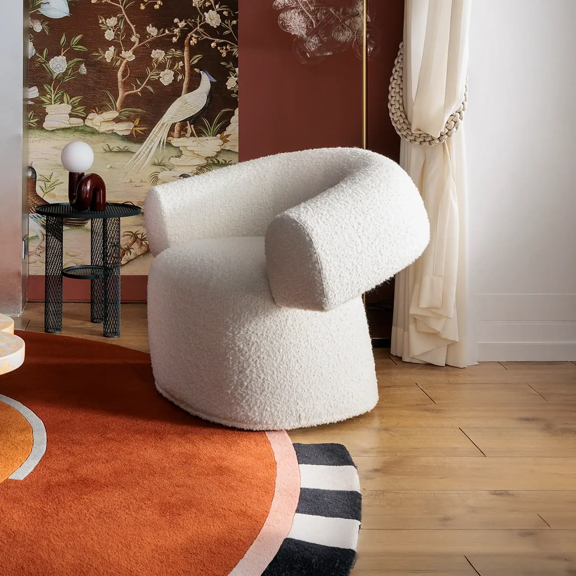 A white boucle armchair in a room with Chinoiserie wallpaper, a black metal side table, and an abstract orange rug.