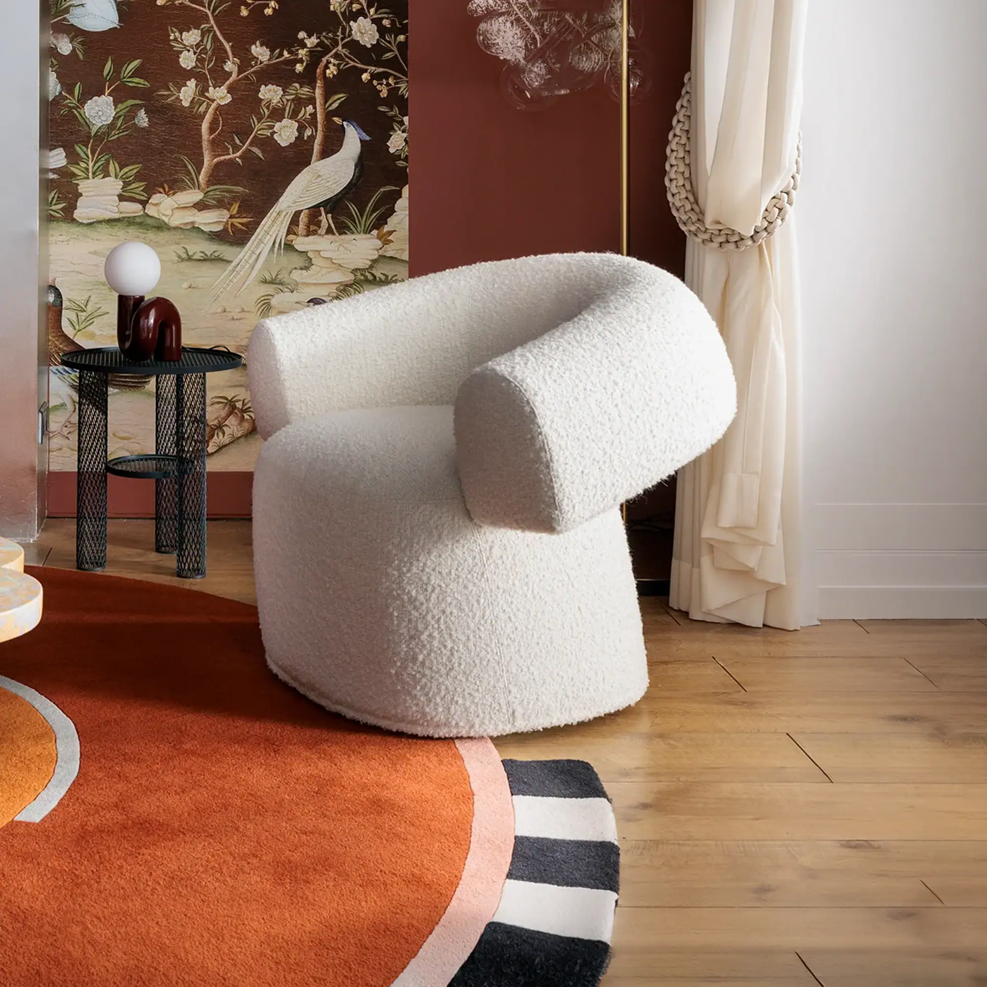 A white boucle armchair in a room with Chinoiserie wallpaper, a black metal side table, and an abstract orange rug.