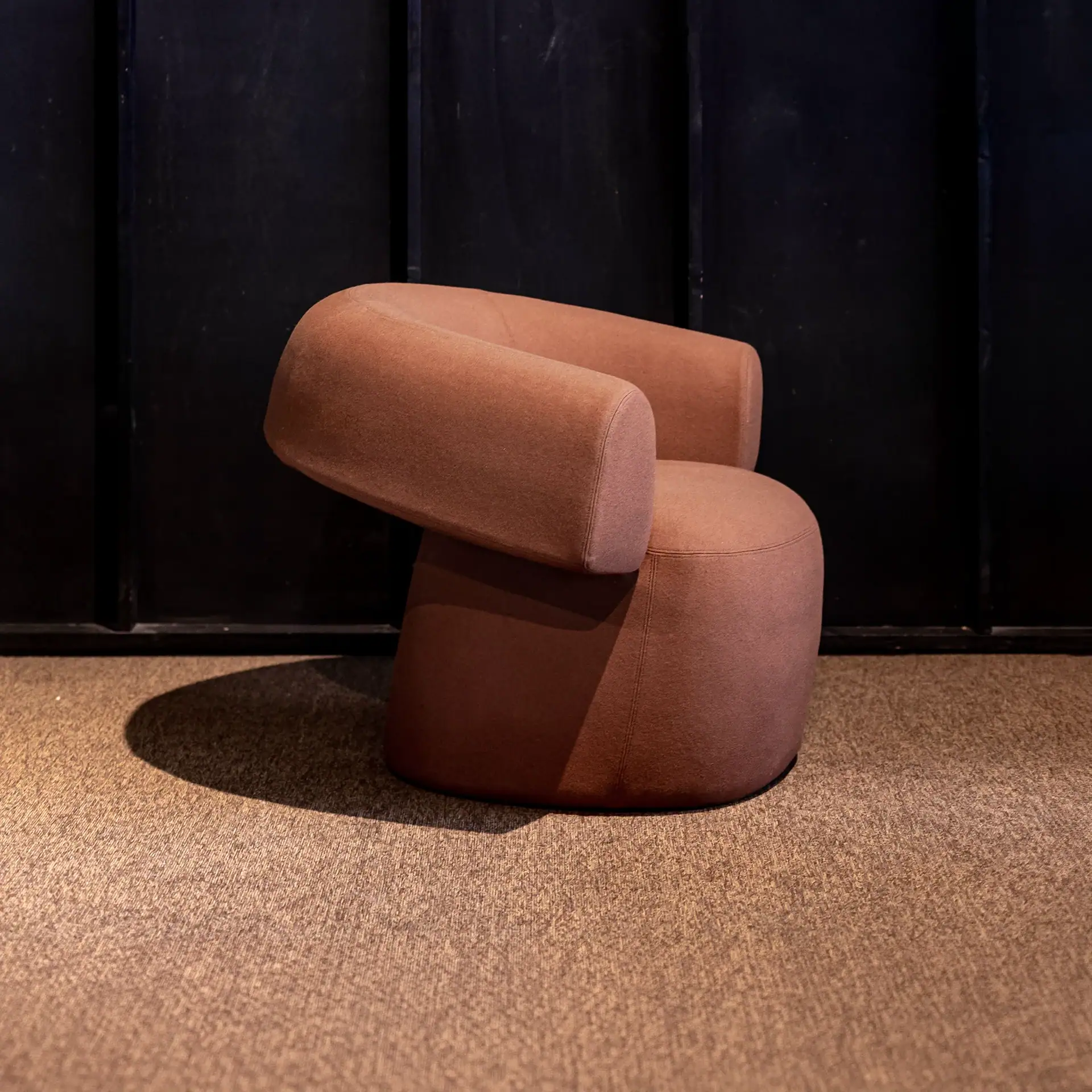A modern, minimalist armchair upholstered in a muted reddish-brown fabric, sitting on a light brown carpet against a dark background.