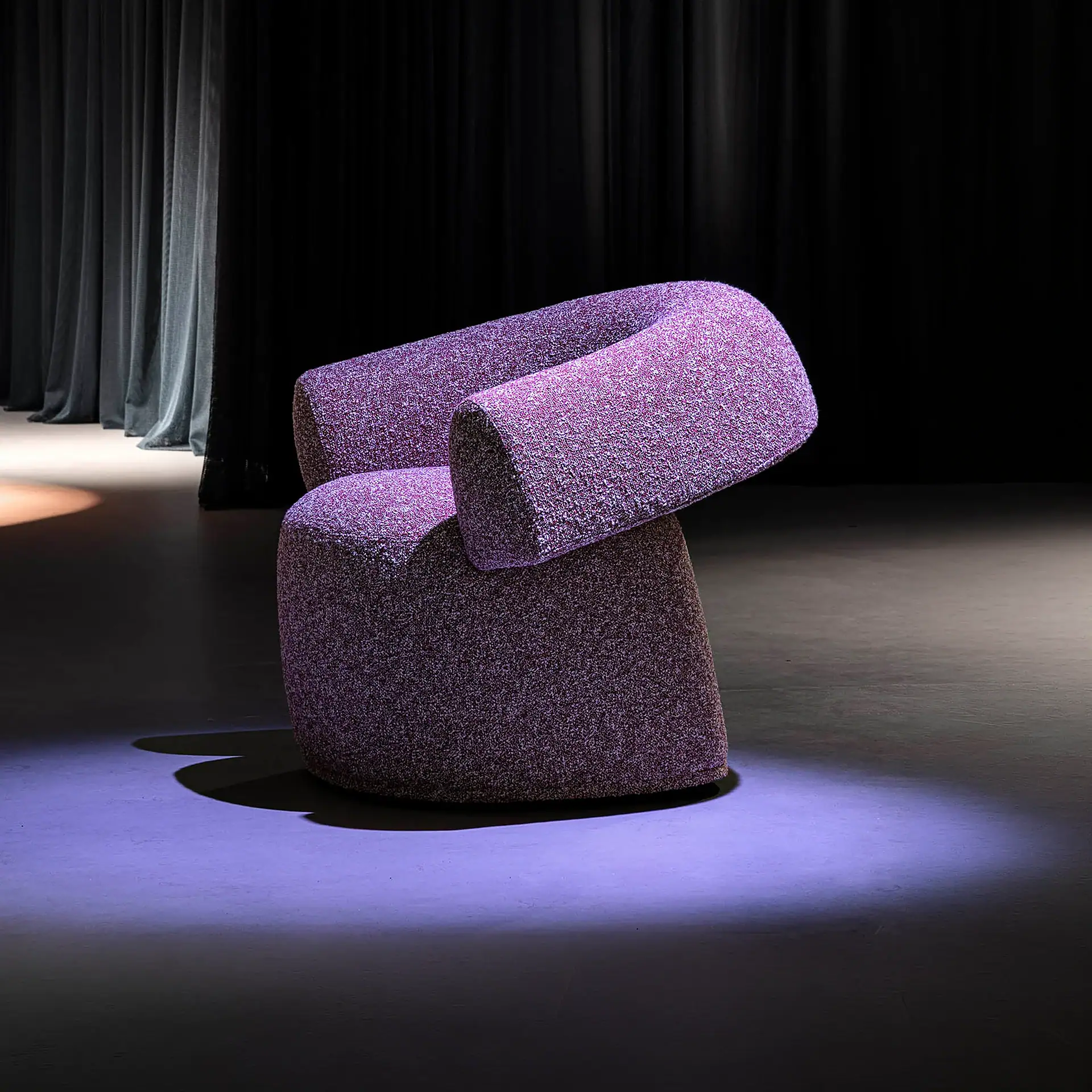 A modern, curved purple armchair with speckled fabric, dramatically lit on a dark stage with curtains in the background.