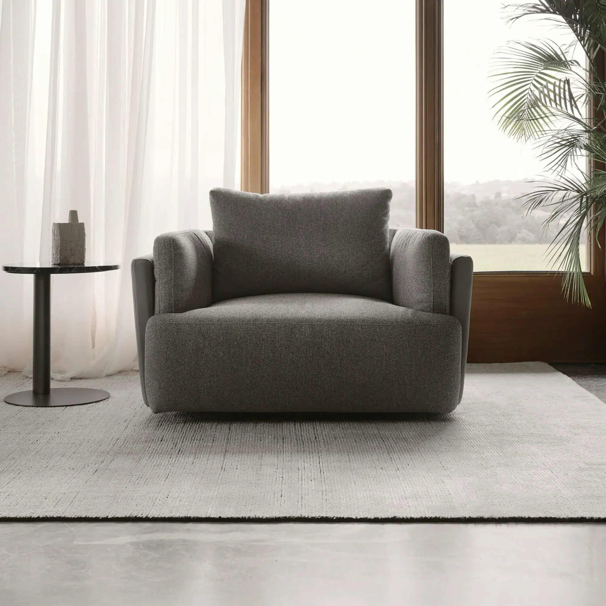 A grey upholstered armchair sits on a light grey rug in front of a large window with a green landscape view. A black side table with a grey vase is to the left, and a green plant is to the right.