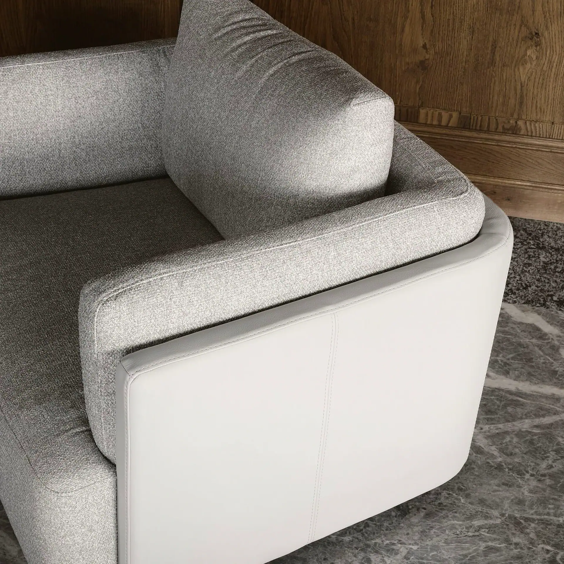 Close-up of a modern armchair with light gray fabric and white leather armrests, on a marble floor next to a wooden wall.