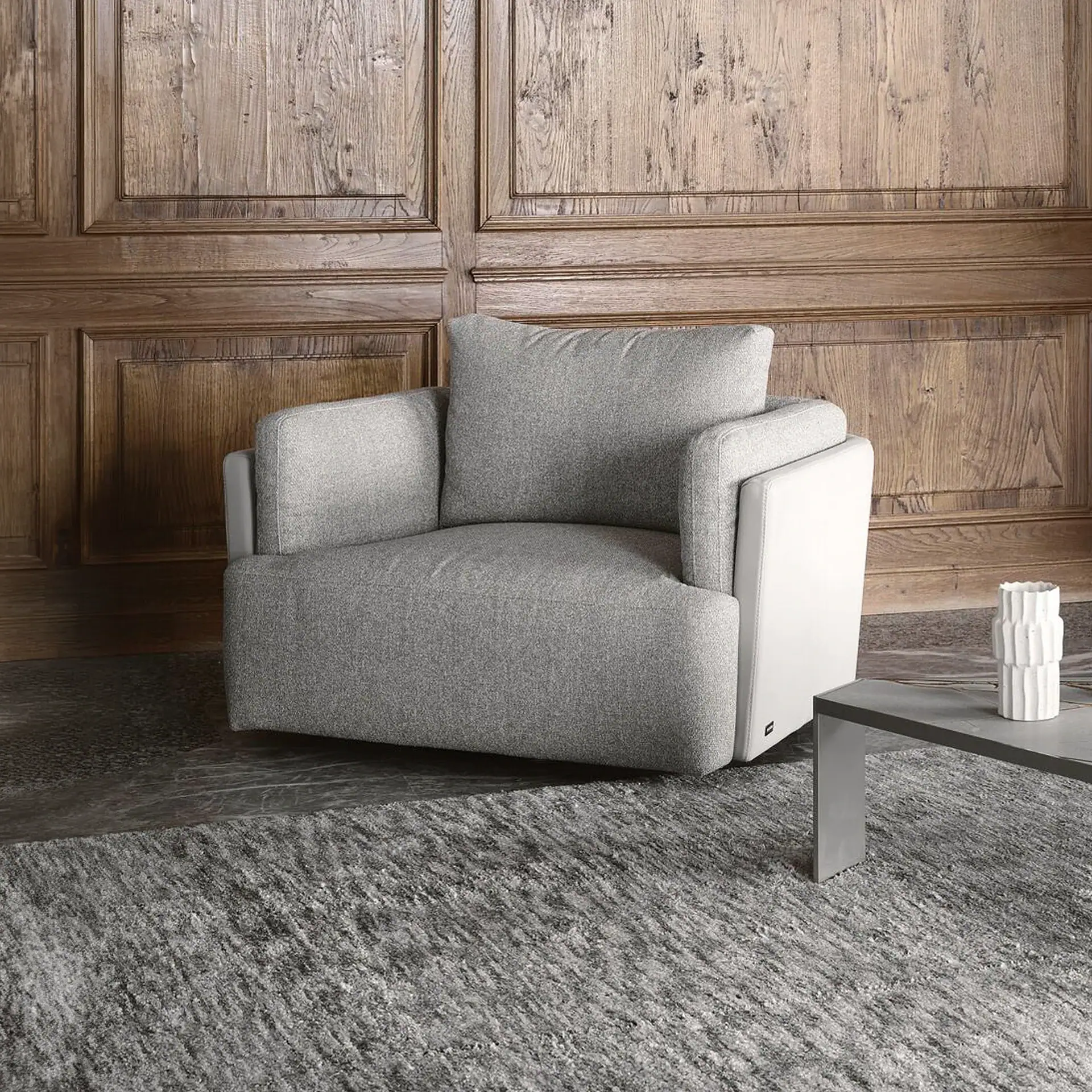 A grey fabric armchair with white side panels sits on a grey rug in front of a dark wooden paneled wall. A white vase is on a grey table to the right.