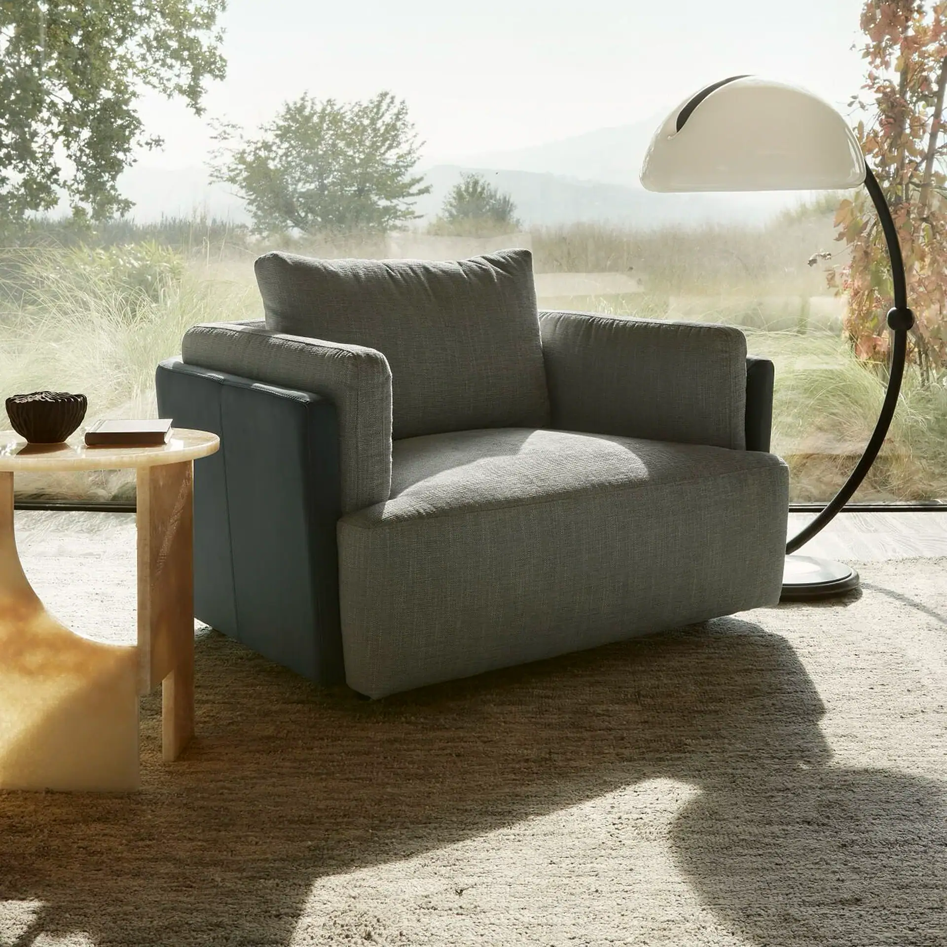 A grey upholstered armchair, a light-colored side table with a decorative bowl and book, and a white floor lamp in a room with a large window overlooking a natural landscape.