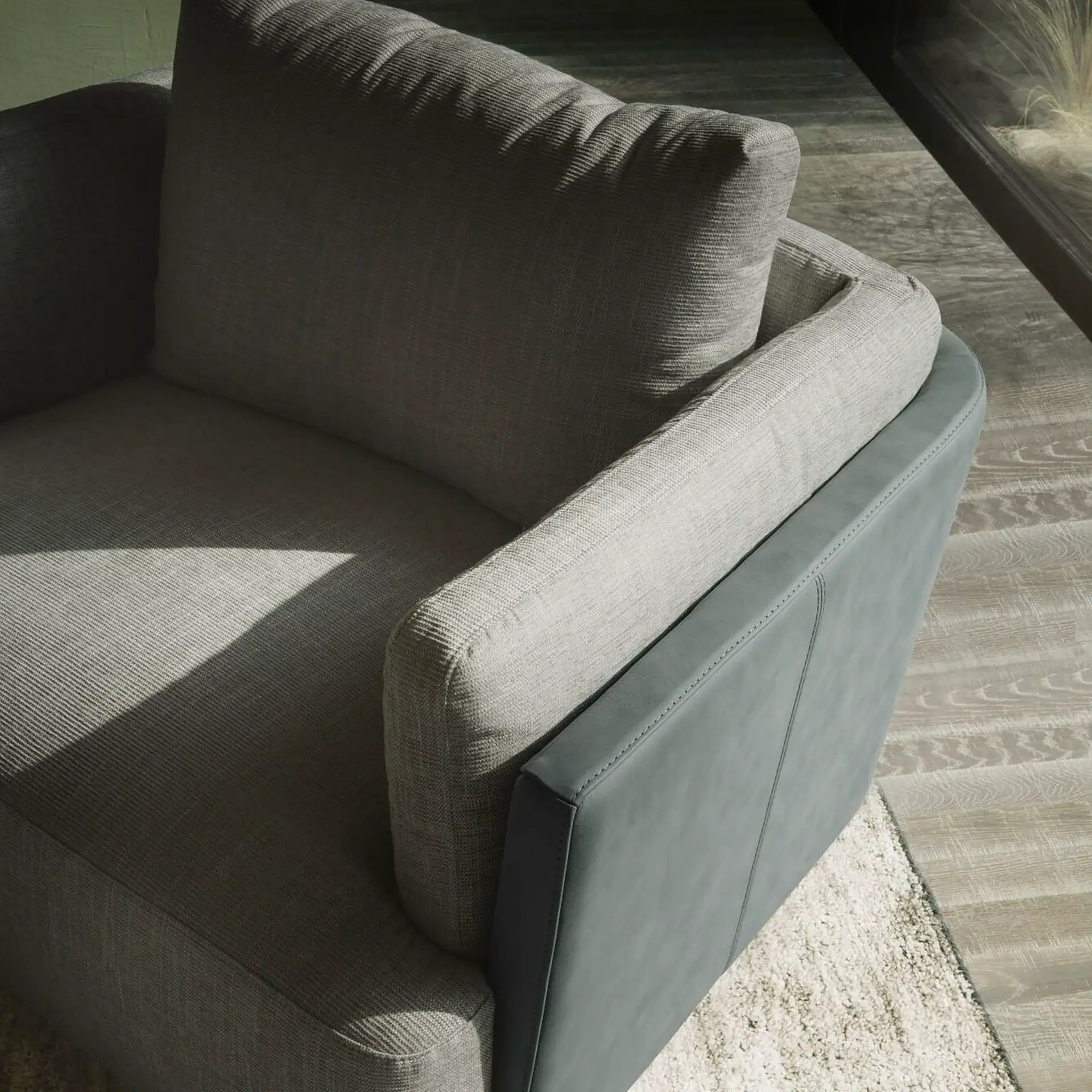 A modern armchair with light grey fabric and dark teal leather accents, casting shadows from natural light, on a rug and wooden floor.