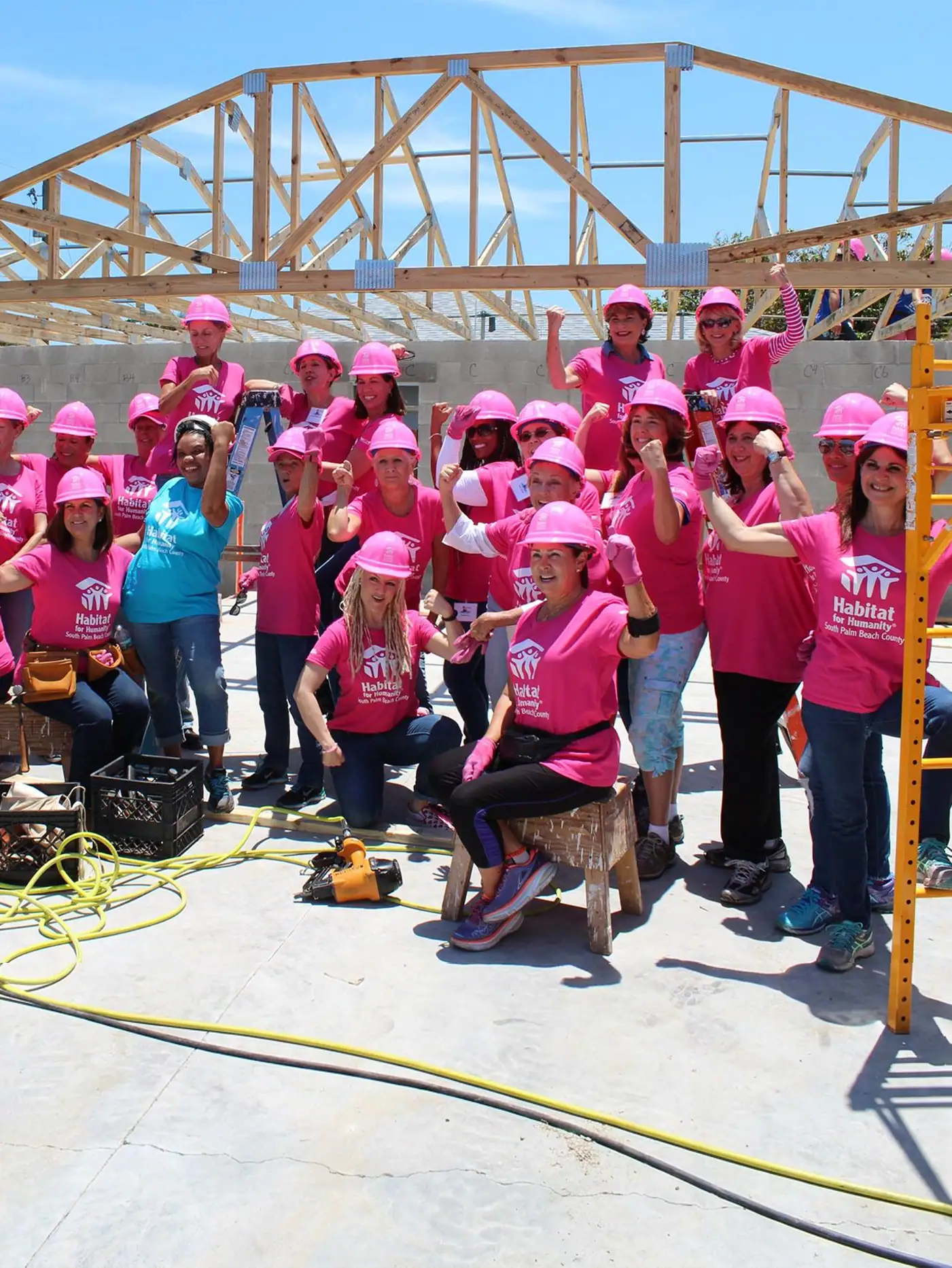 A large group of Habitat for Humanity volunteers, mostly women in pink t-shirts and hard hats, flexing their muscles at a construction site with a partially built house in the background.