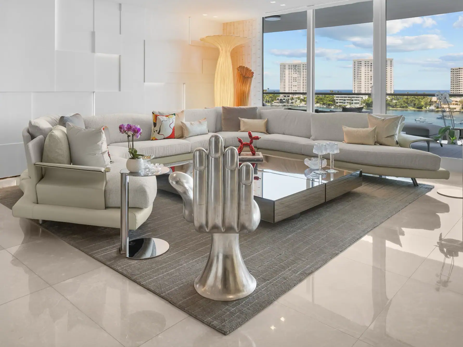 our core values encourage creativity 1.4 | Sklar Furnishings A modern living room with a large sectional sofa, a mirrored coffee table, a silver hand sculpture, and floor-to-ceiling windows overlooking the ocean and city skyline.