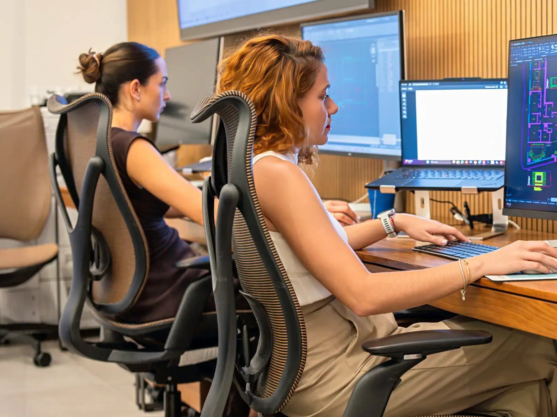 A woman with curly reddish-brown hair works at a desk with multiple computer monitors, one displaying architectural plans, and a laptop on a stand.