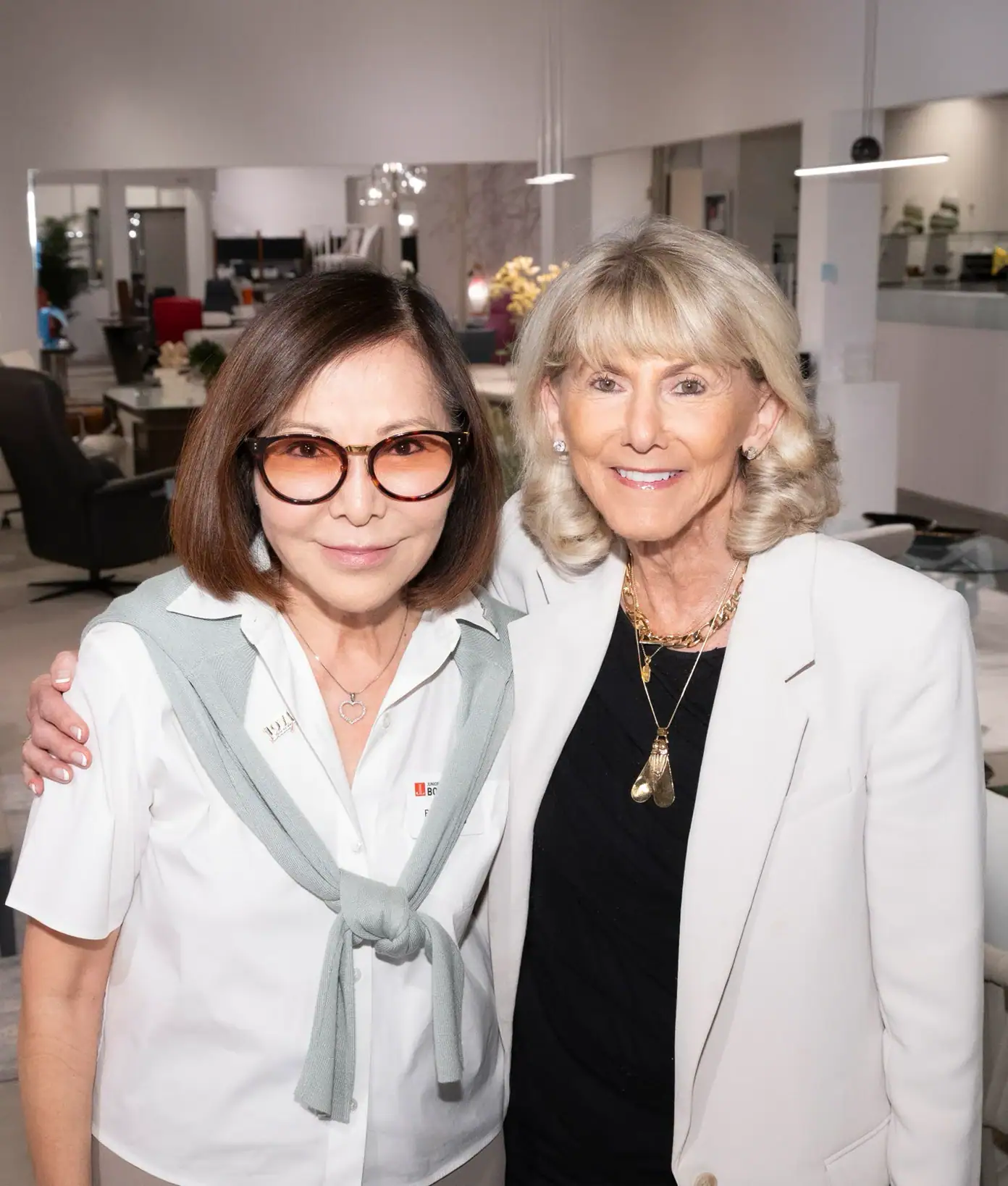our core values inspire | Sklar Furnishings Two smiling women, one with dark hair and glasses, the other with blonde hair, pose together in a modern furniture showroom.