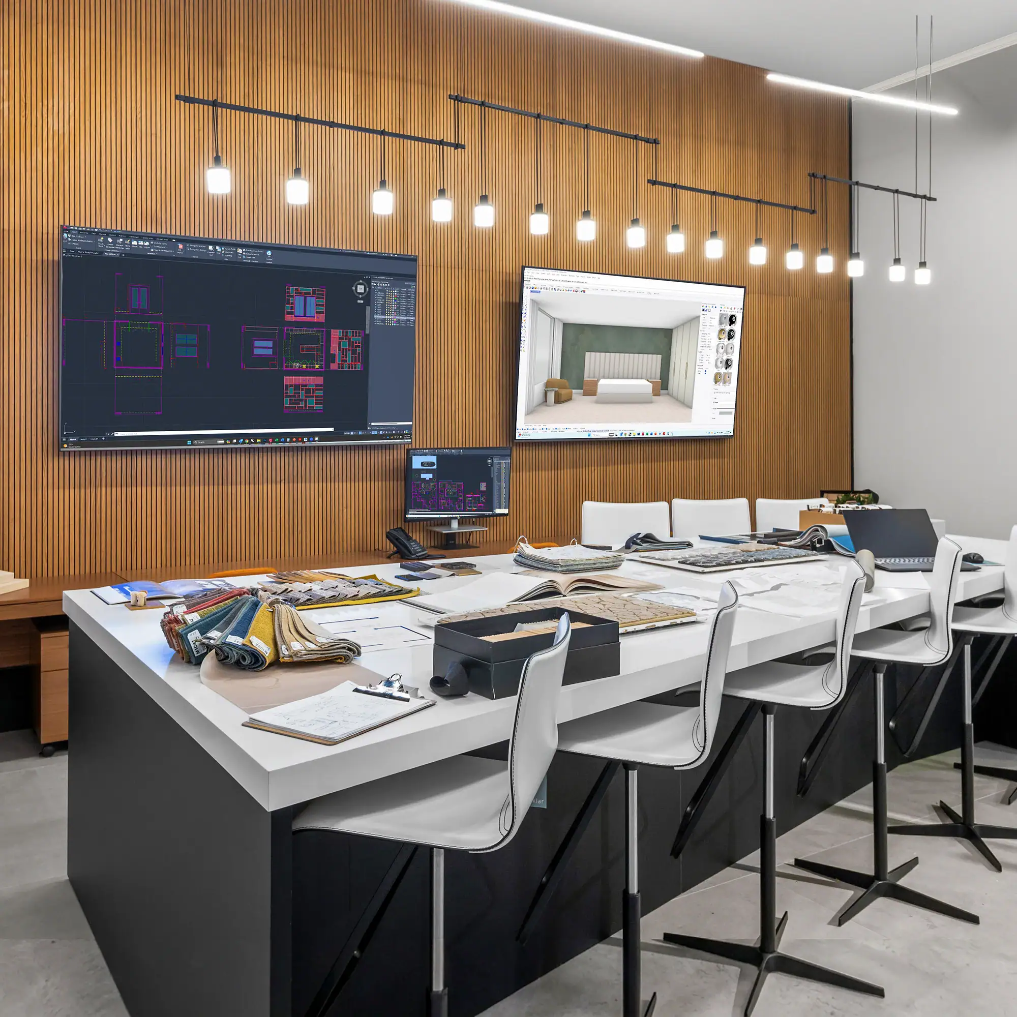 A modern design studio with a long white table covered in fabric swatches and design materials, two large screens displaying architectural plans and 3D interior renderings, and white bar stools.