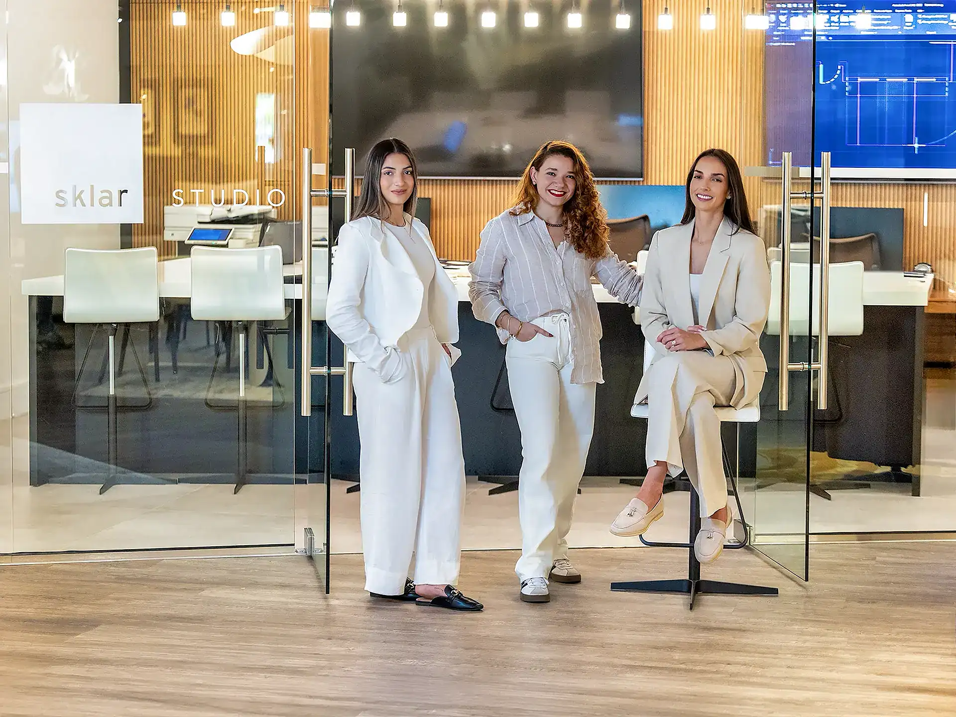 Three women smiling and posing in front of a glass-walled office with 'sklar STUDIO' signage. They are dressed in light-colored business casual attire.