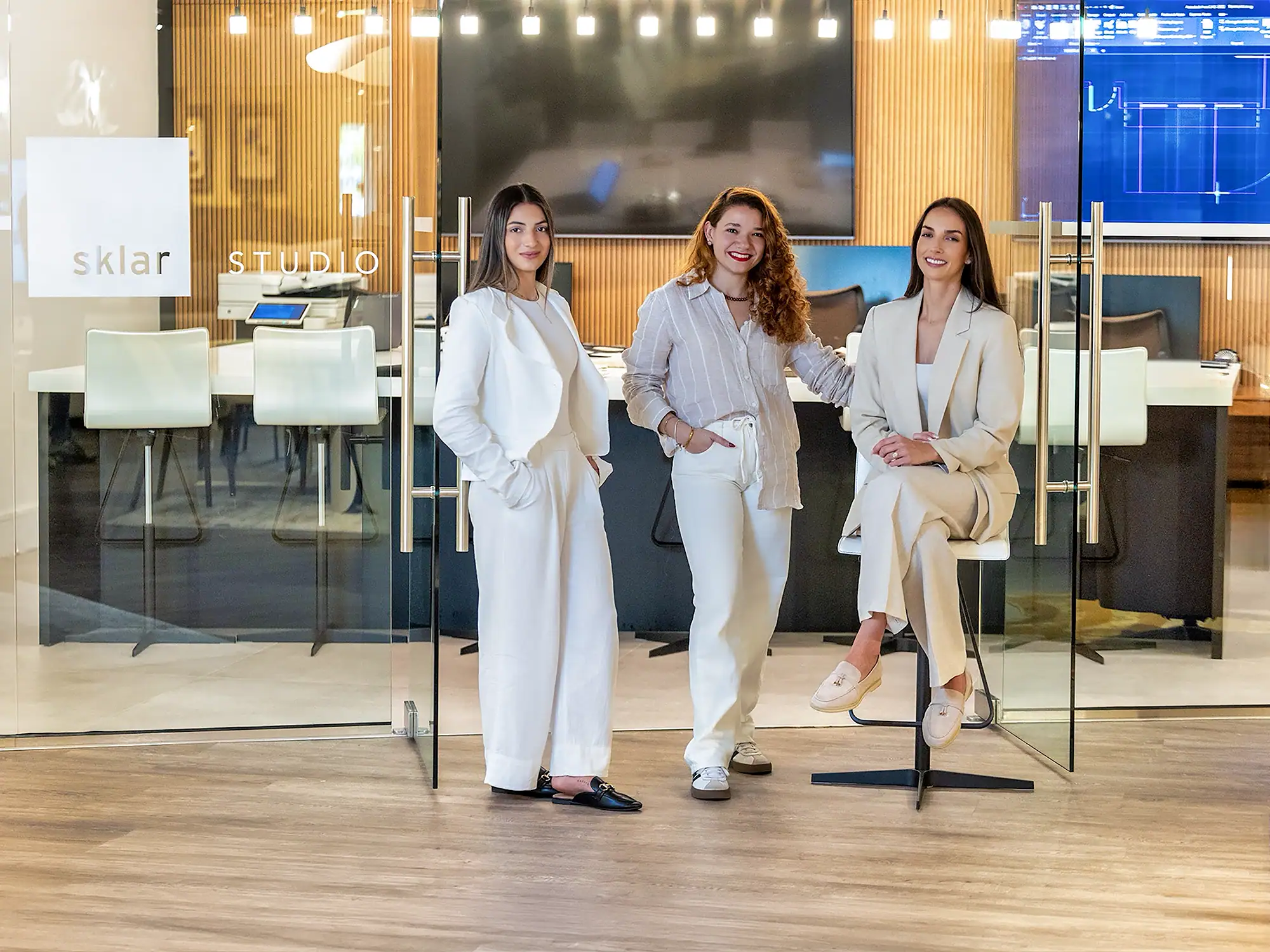 Three women smiling and posing in front of a glass-walled office with 'sklar STUDIO' signage. They are dressed in light-colored business casual attire.