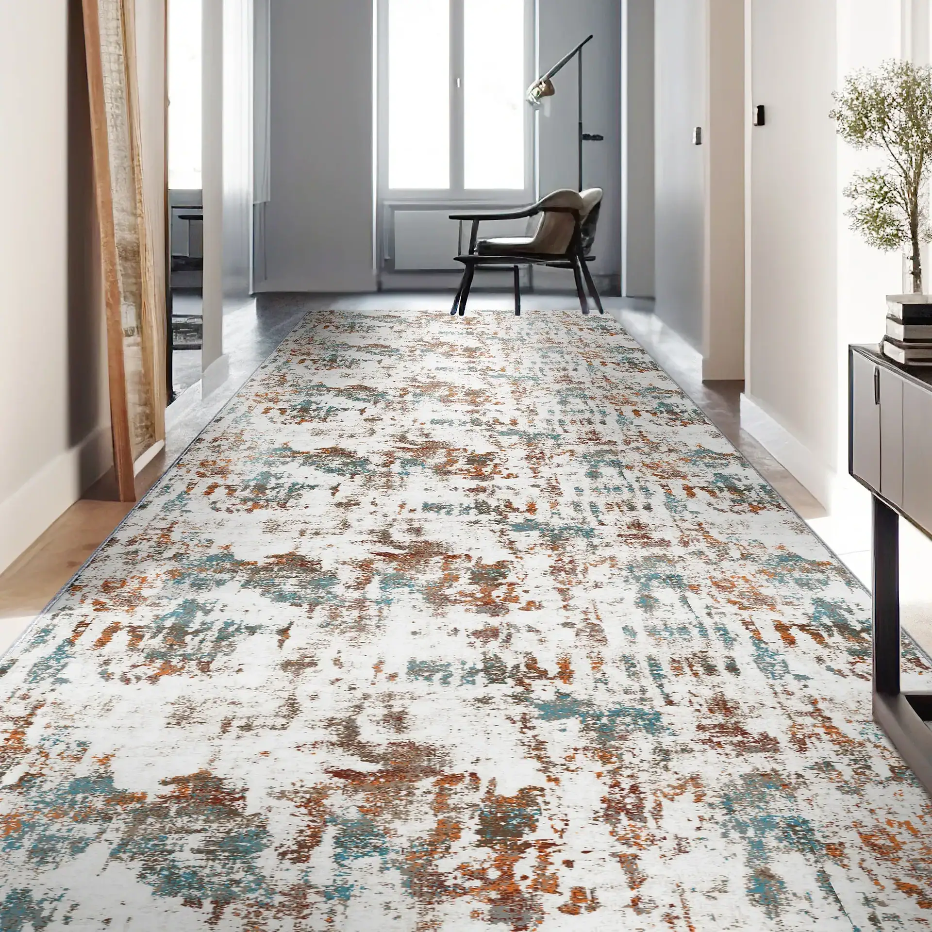 A long hallway with a white, teal, and rust abstract runner rug. A mirror is on the left, a console table with a plant on the right, and an armchair by a window at the end.