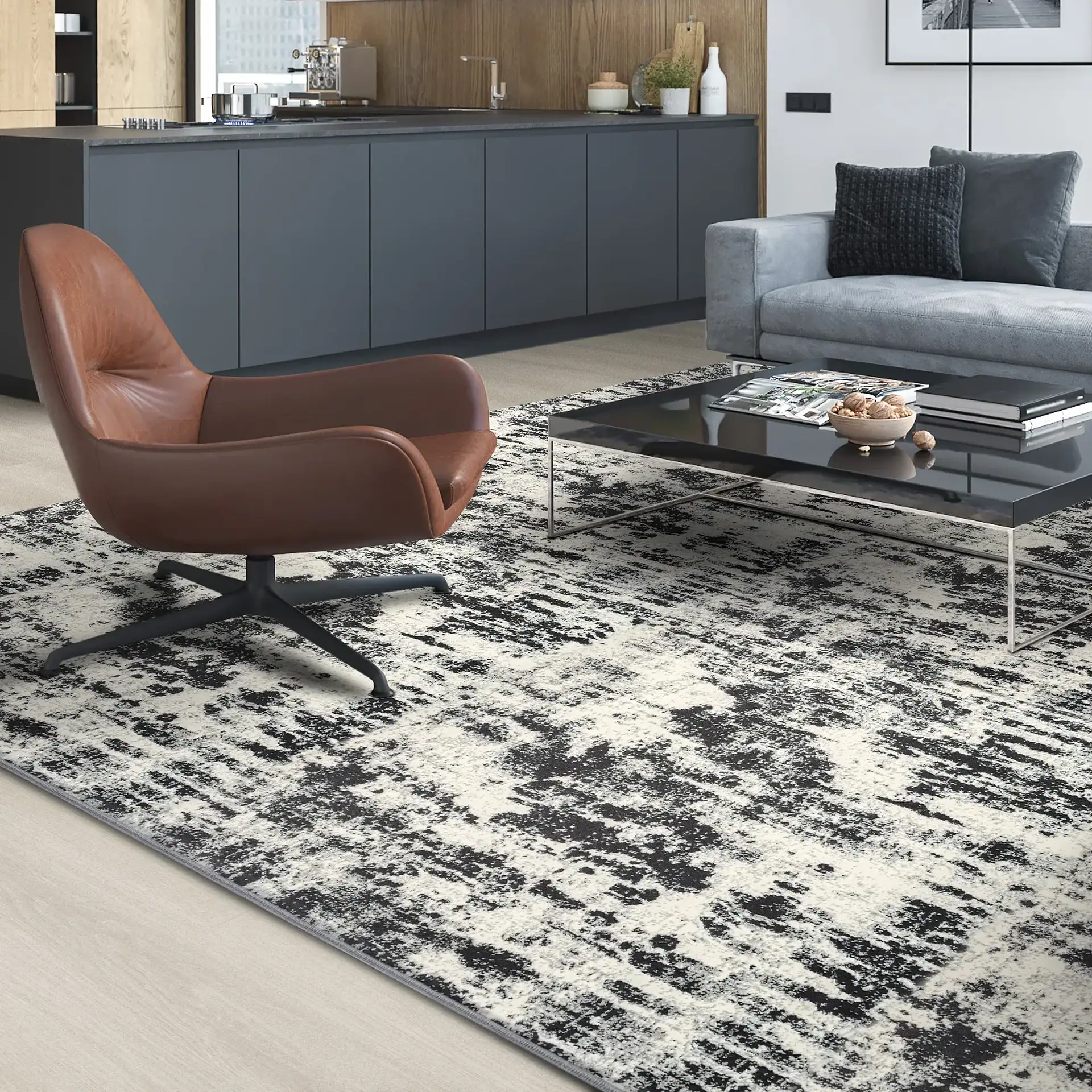 A modern living room and kitchen area with a black and white abstract rug, a brown leather armchair, a gray sofa, and a black coffee table.