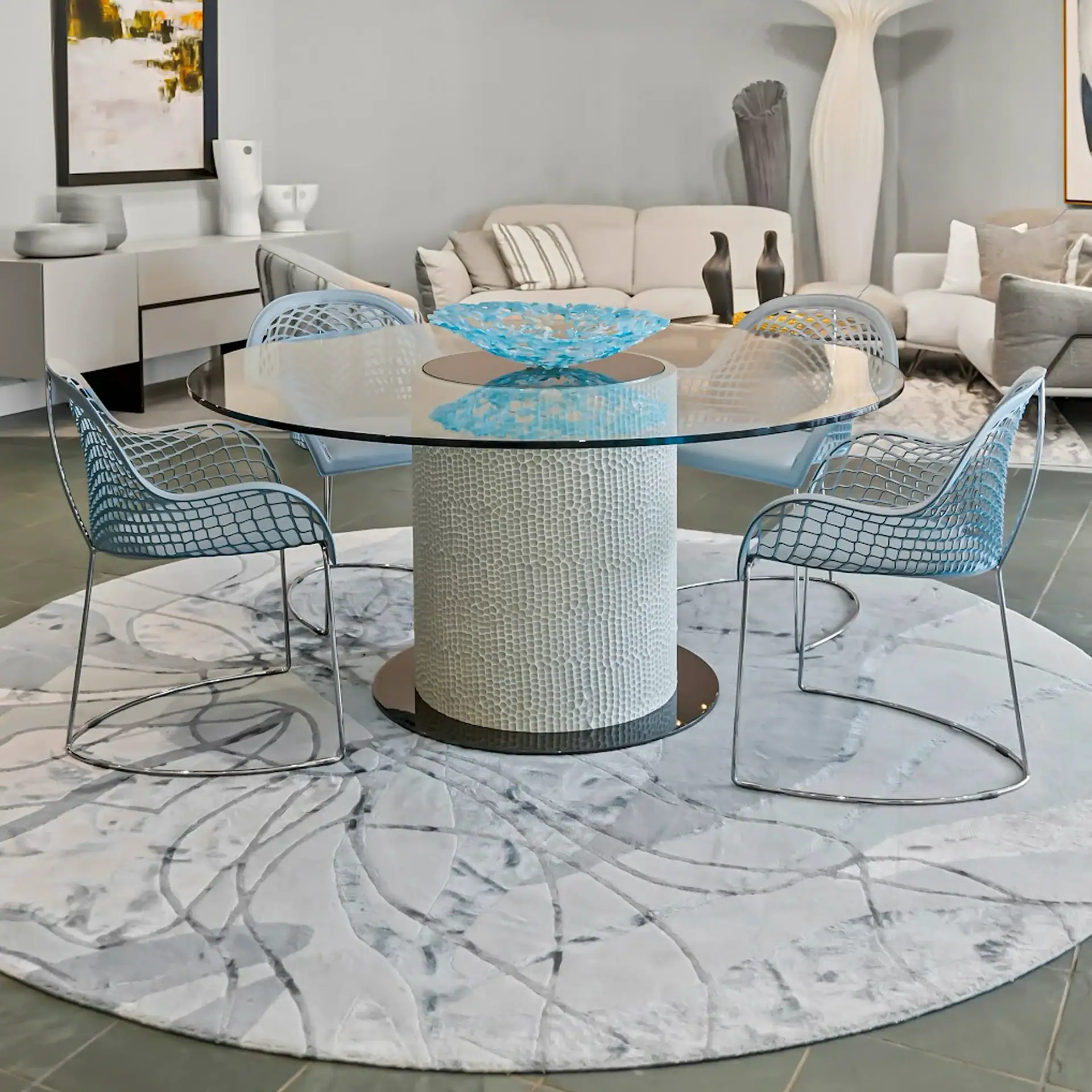 A modern dining room with a round glass table, blue mesh chairs, and a blue glass bowl on an abstract rug. A living room with a sofa and abstract art is in the background.