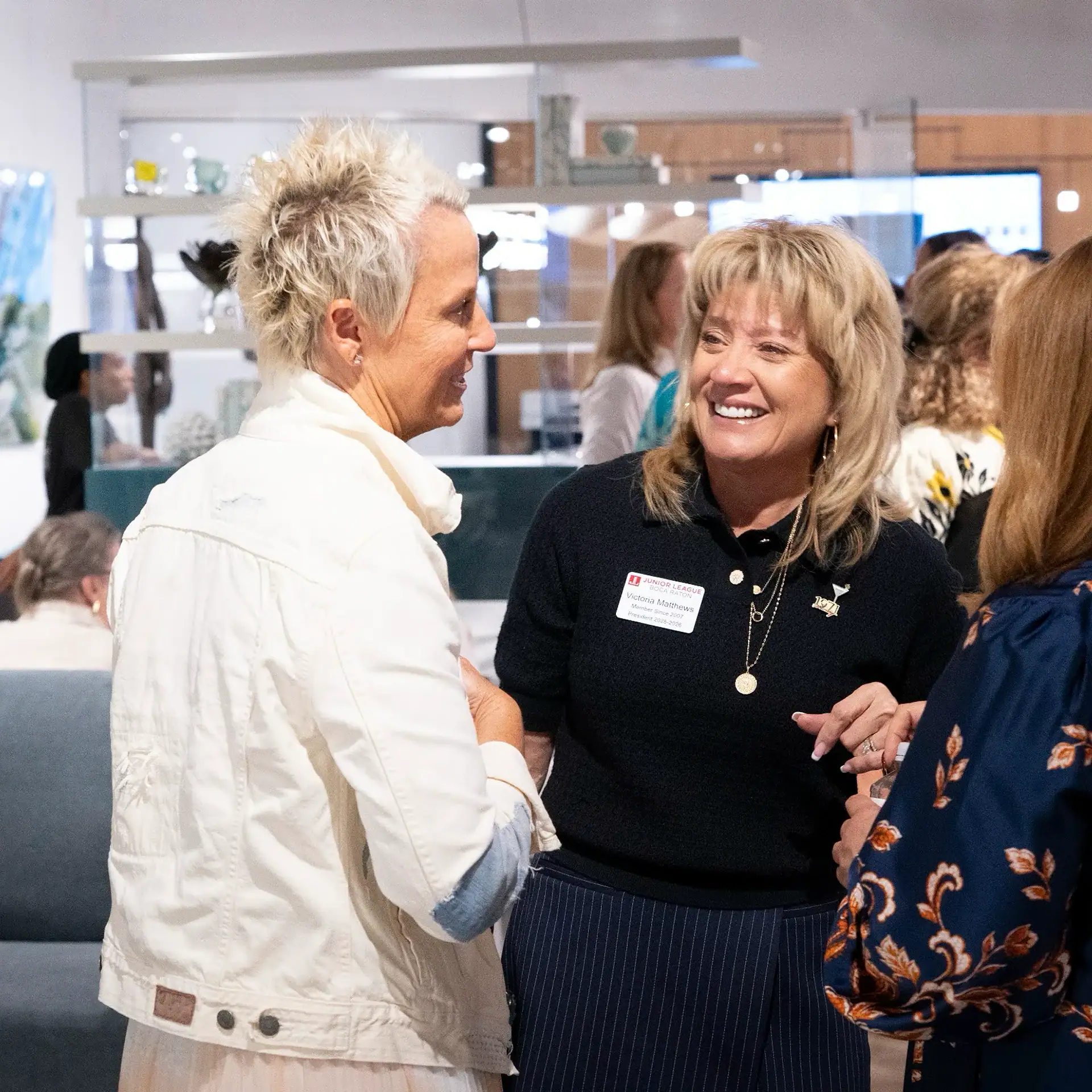 Two women smiling and talking at an indoor event. One woman wears a name tag for the Junior League of Boca Raton.