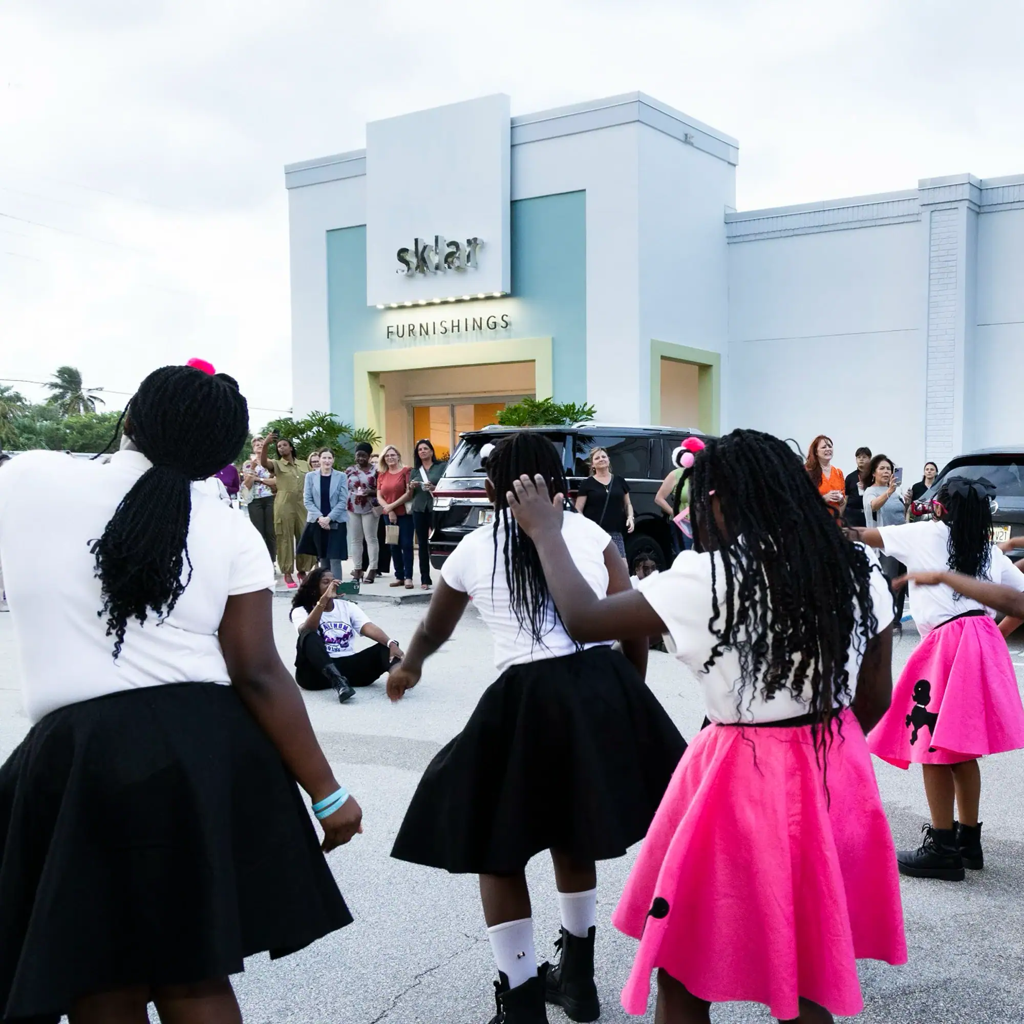 the-sklar-experience-our-commitment-1 Several young Black girls in white t-shirts and 1950s-style poodle skirts dance outdoors in front of a building, with adults watching.