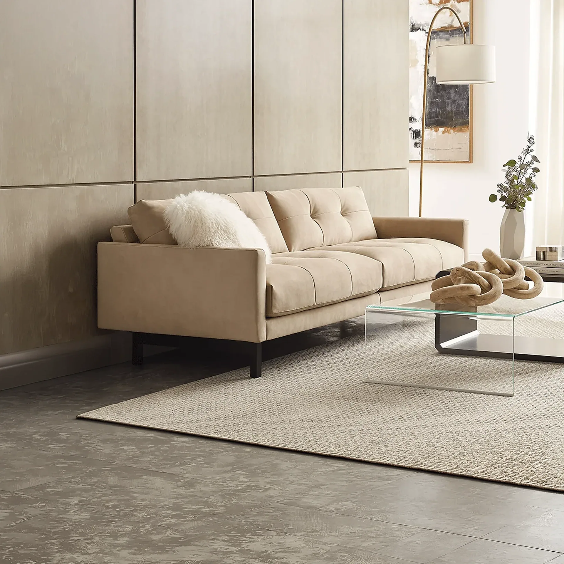 A modern living room with a beige tufted sofa, glass coffee table, and light rug on a dark tiled floor. A brass floor lamp and abstract art are visible in the background.