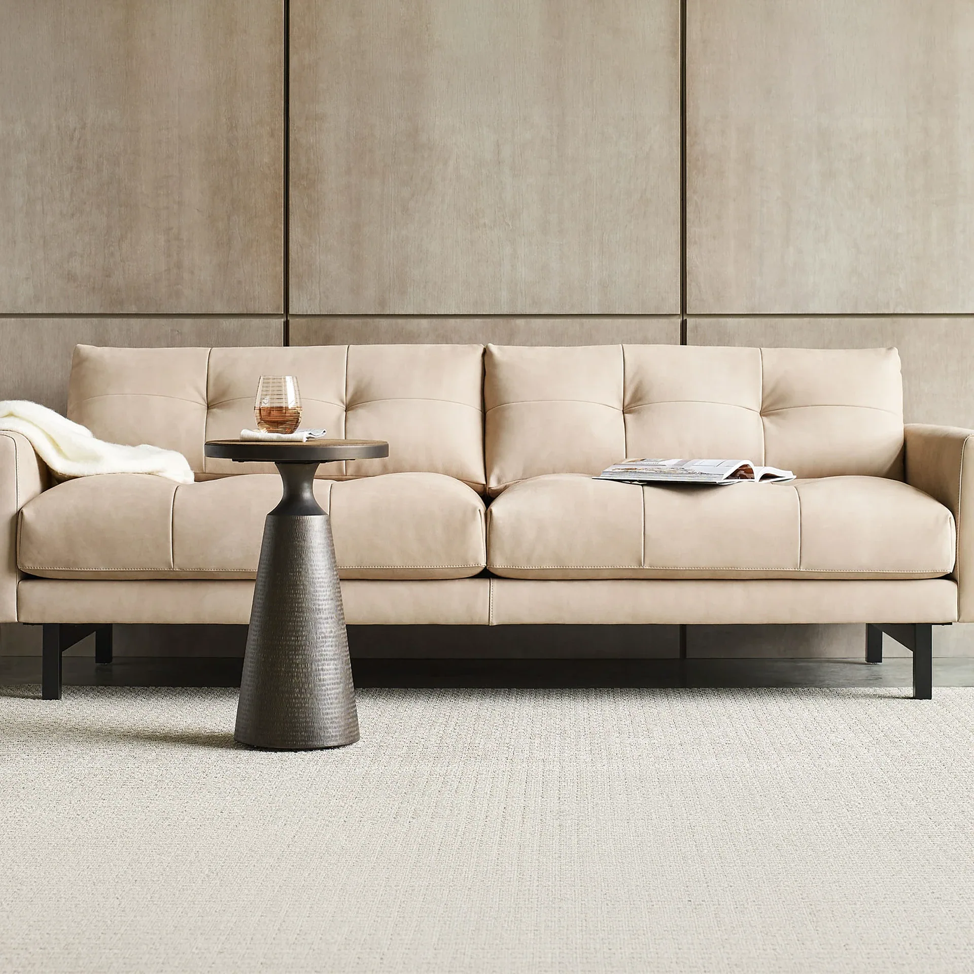A light beige tufted leather sofa with black legs, a dark bronze side table with a drink, and a magazine, all on a light textured rug against a paneled wall.