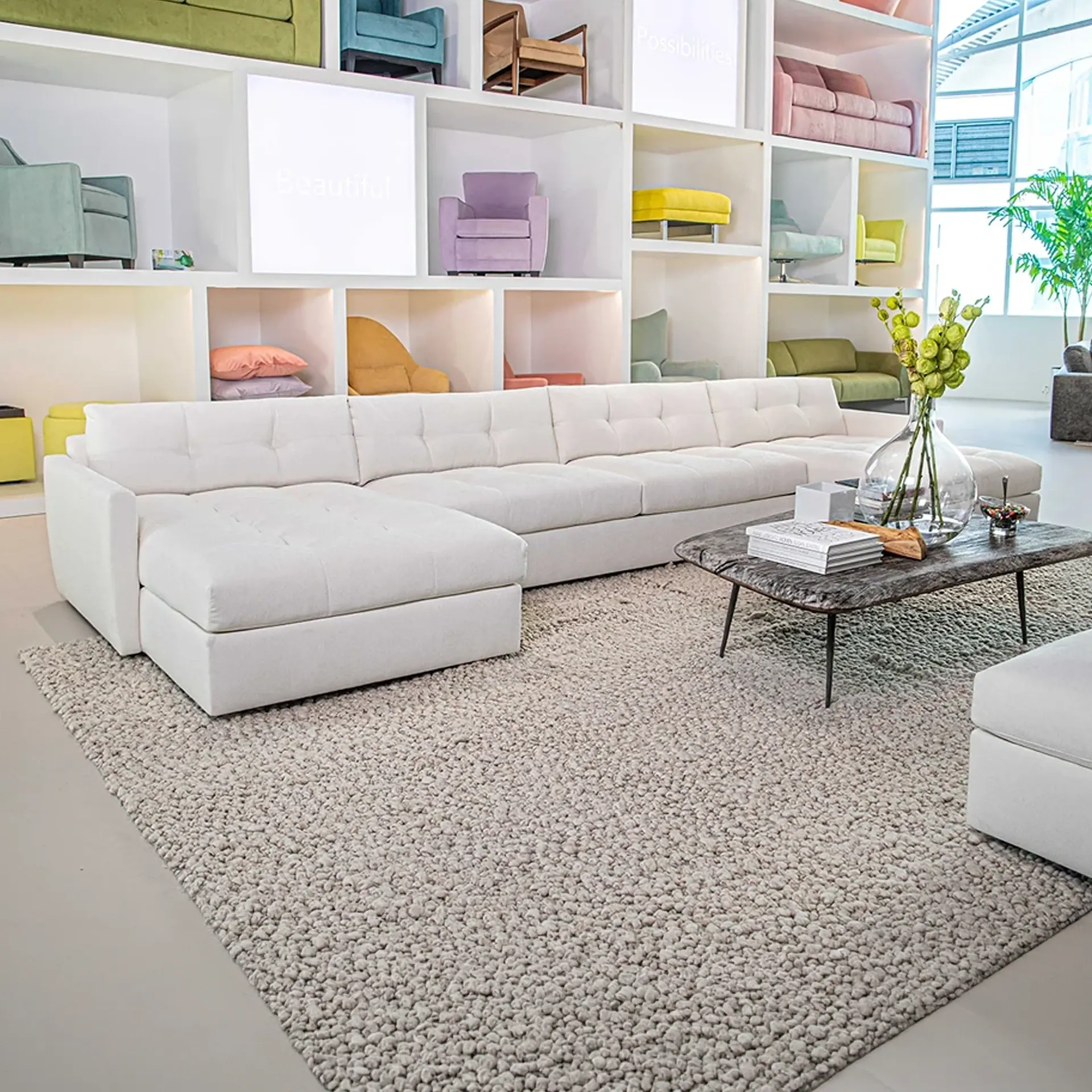 A modern living room display with a large white sectional sofa, a dark gray coffee table, and a textured gray rug. The background shows a white shelving unit filled with colorful miniature furniture displays and text panels reading 'Beautiful' and 'Possibilities'.