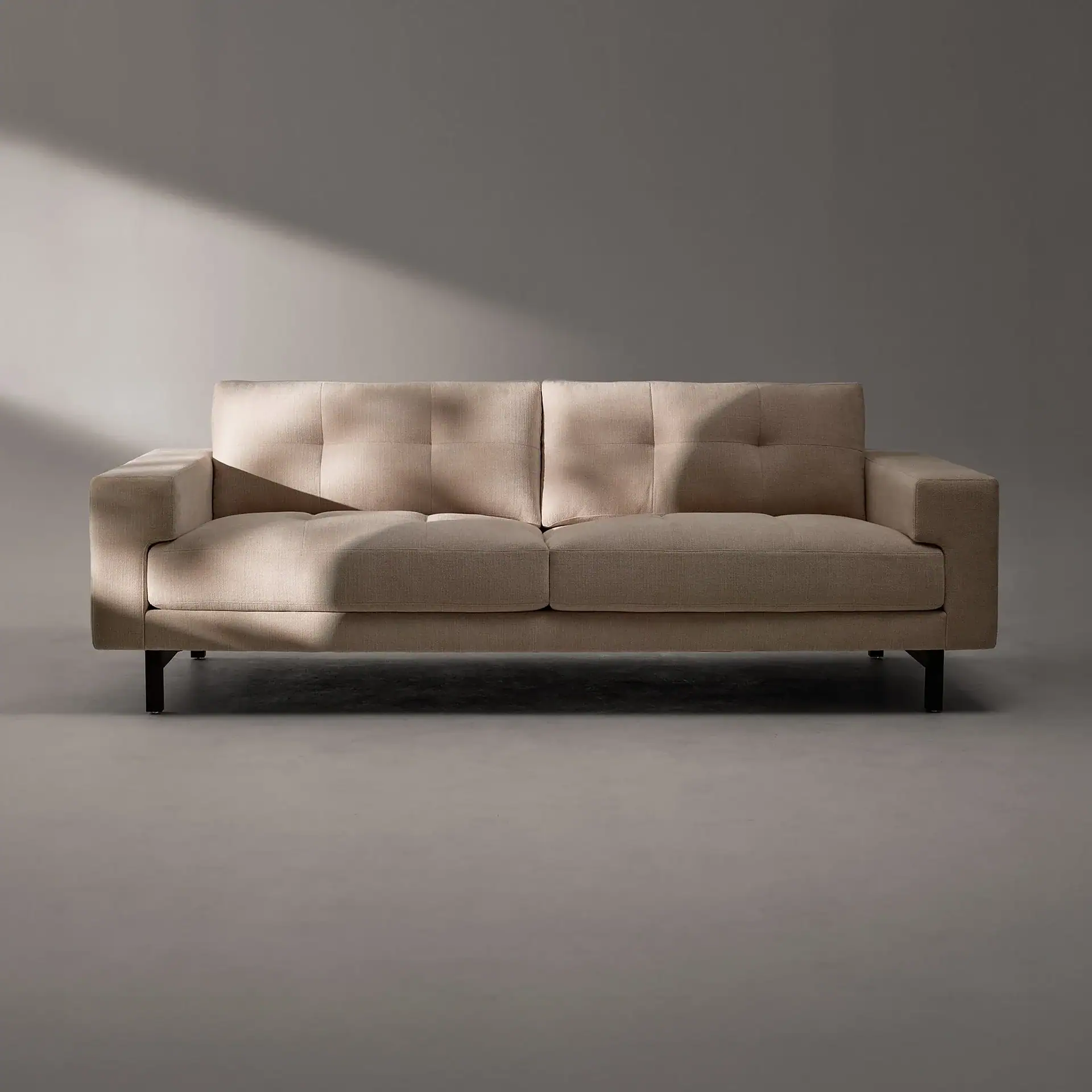 A light beige two-seater sofa with tufted back cushions and dark legs, set against a gray background with a diagonal shadow cast across it.