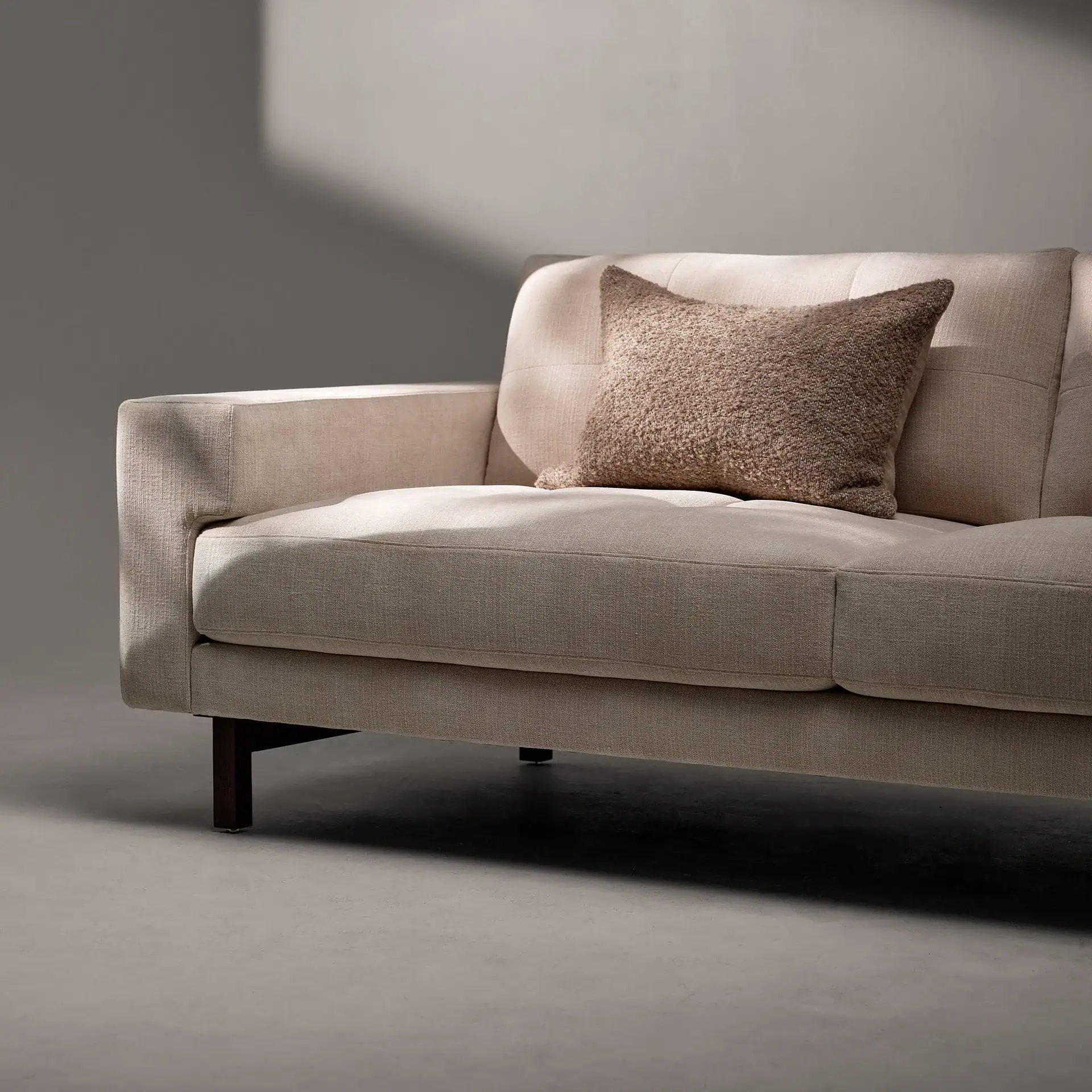 A modern, light beige fabric sofa with a textured light brown throw pillow, illuminated by natural light casting shadows on the wall and sofa.