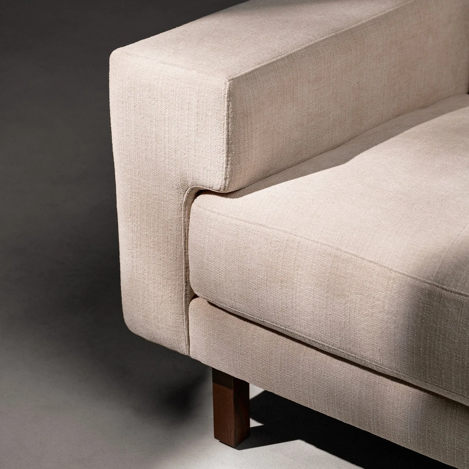Close-up of a light beige fabric sofa armrest and seat cushion with a dark wooden leg, casting shadows on a gray background.