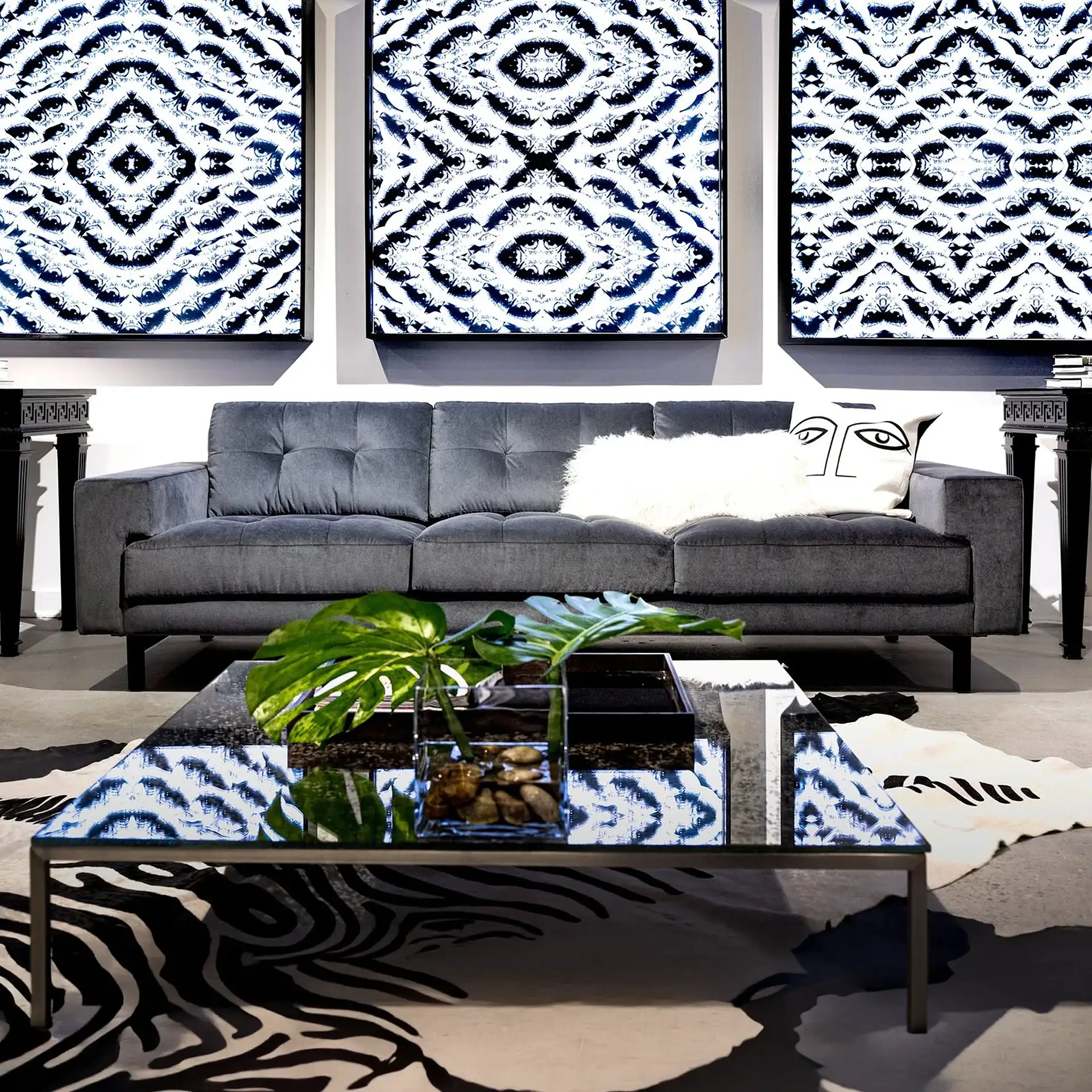 A modern living room with a dark gray sofa, three framed blue and white abstract eye-pattern artworks, a glass coffee table, and zebra print and cowhide rugs on a concrete floor.