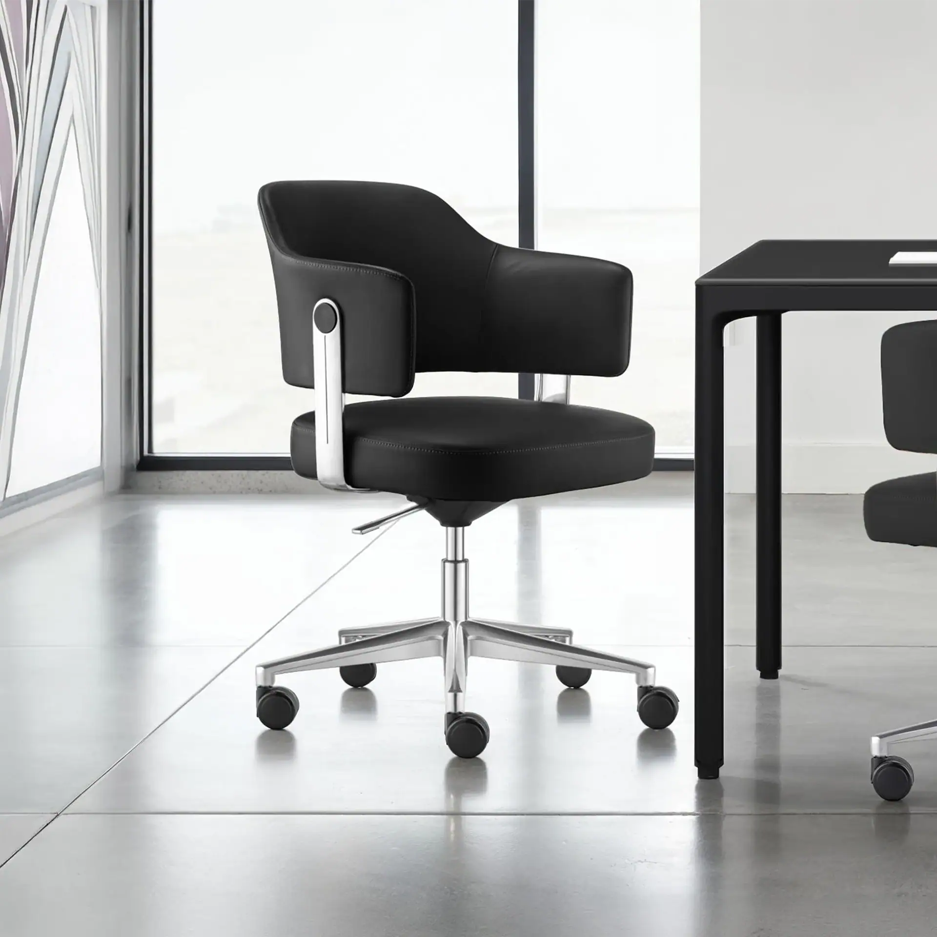 A black leather office chair with chrome accents and a five-star caster base sits on a light grey floor in a modern room with large windows.