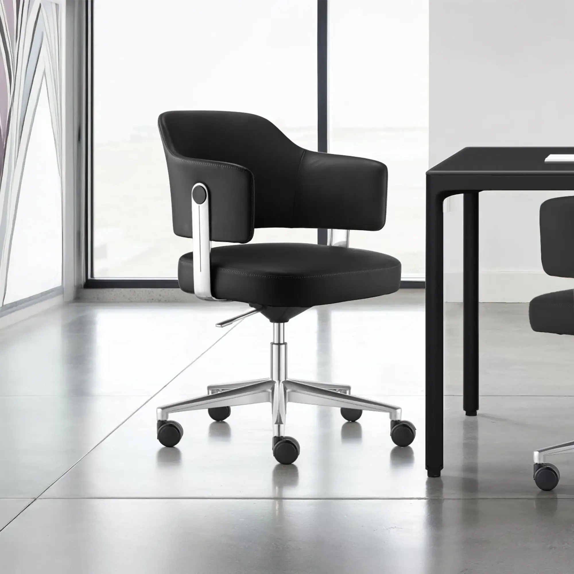 A black leather office chair with chrome accents and a five-star caster base sits on a light grey floor in a modern room with large windows.