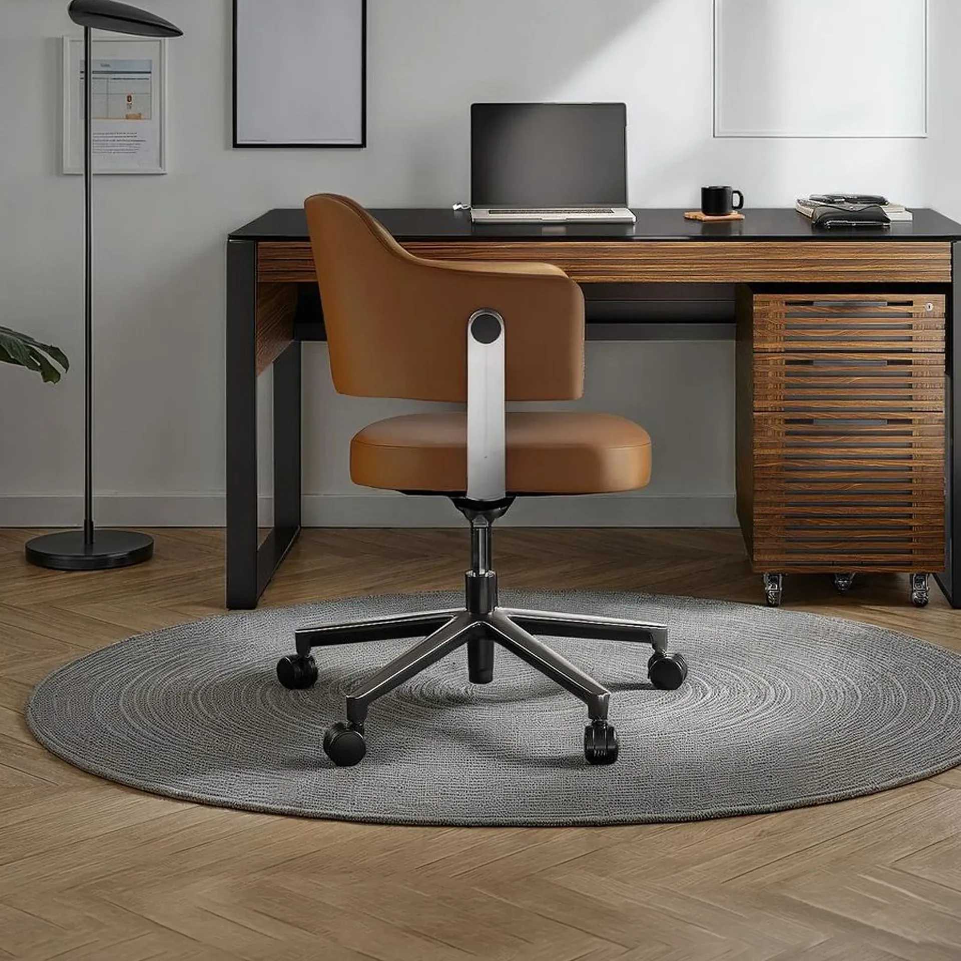 A modern home office with a tan leather office chair on a grey round rug, a dark wooden desk with a laptop, and a black floor lamp.