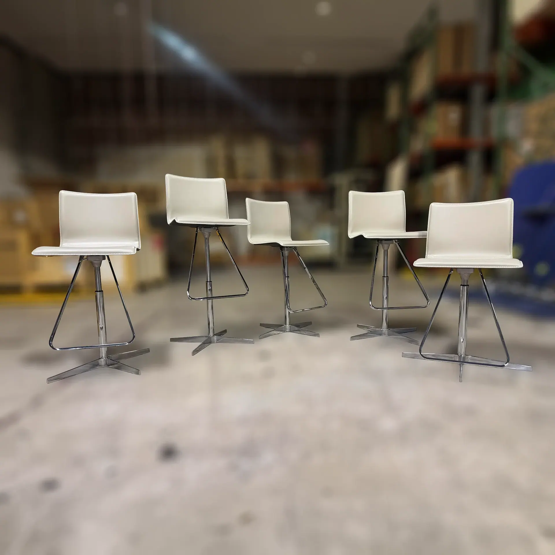 Five modern cream bar stools with chrome bases arranged in a slight curve on a light gray floor, with a blurred warehouse background.