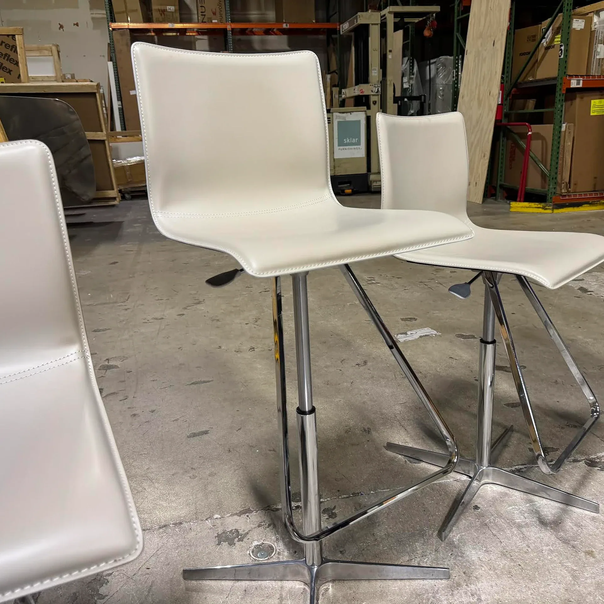Three modern light beige bar stools with chrome bases on a concrete floor in a warehouse setting.