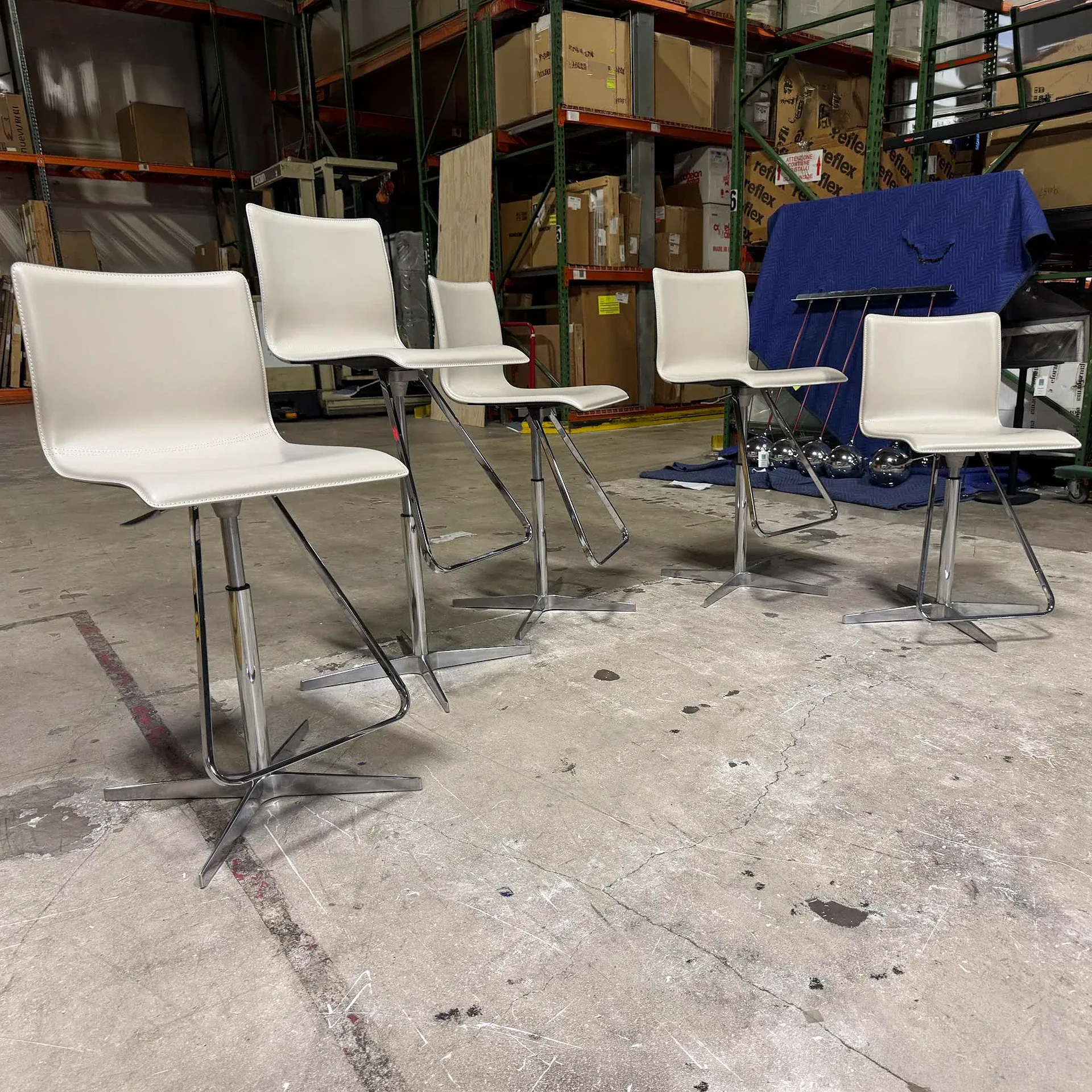 Five white bar stools with chrome bases are lined up on a concrete floor in a warehouse with shelving and boxes in the background.