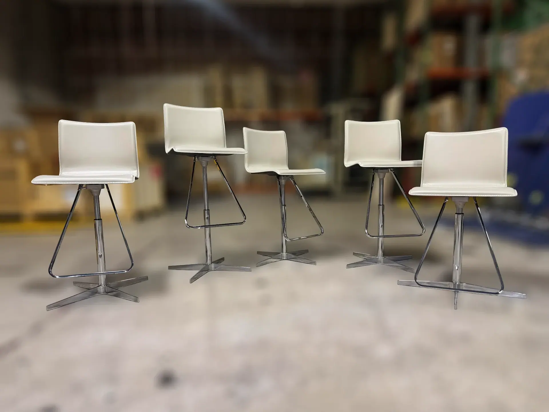 Five cream upholstered bar stools with chrome bases and triangular footrests are arranged in a line against a blurred warehouse background.